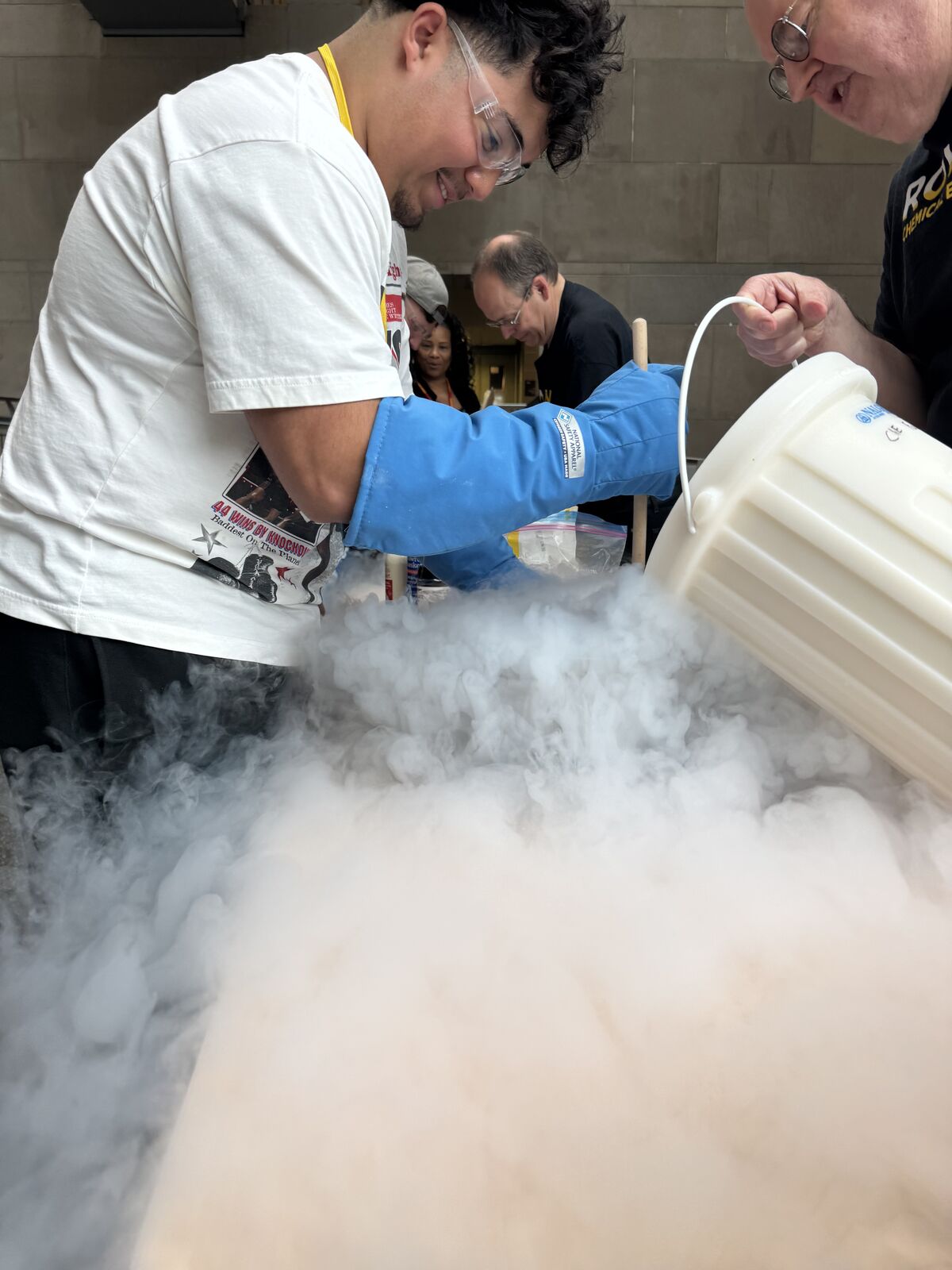 A young person in safety glasses and blue cryogenic gloves pours a white substance from a bucket, creating a large cloud of vapor.