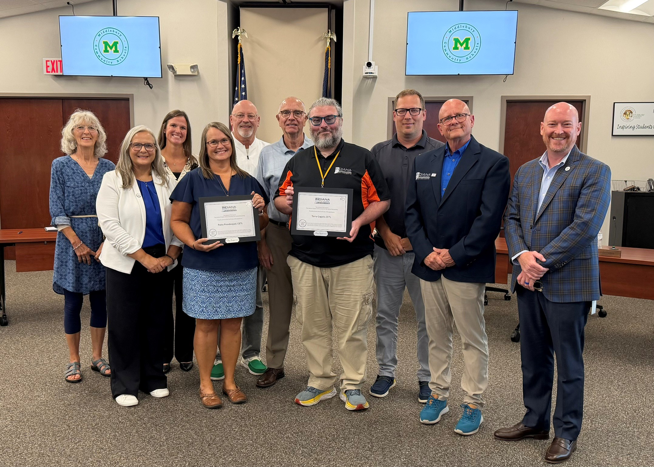 A group of people stand in a room, some holding certificates, with a "Middlebury Community Schools" logo displayed on screens.