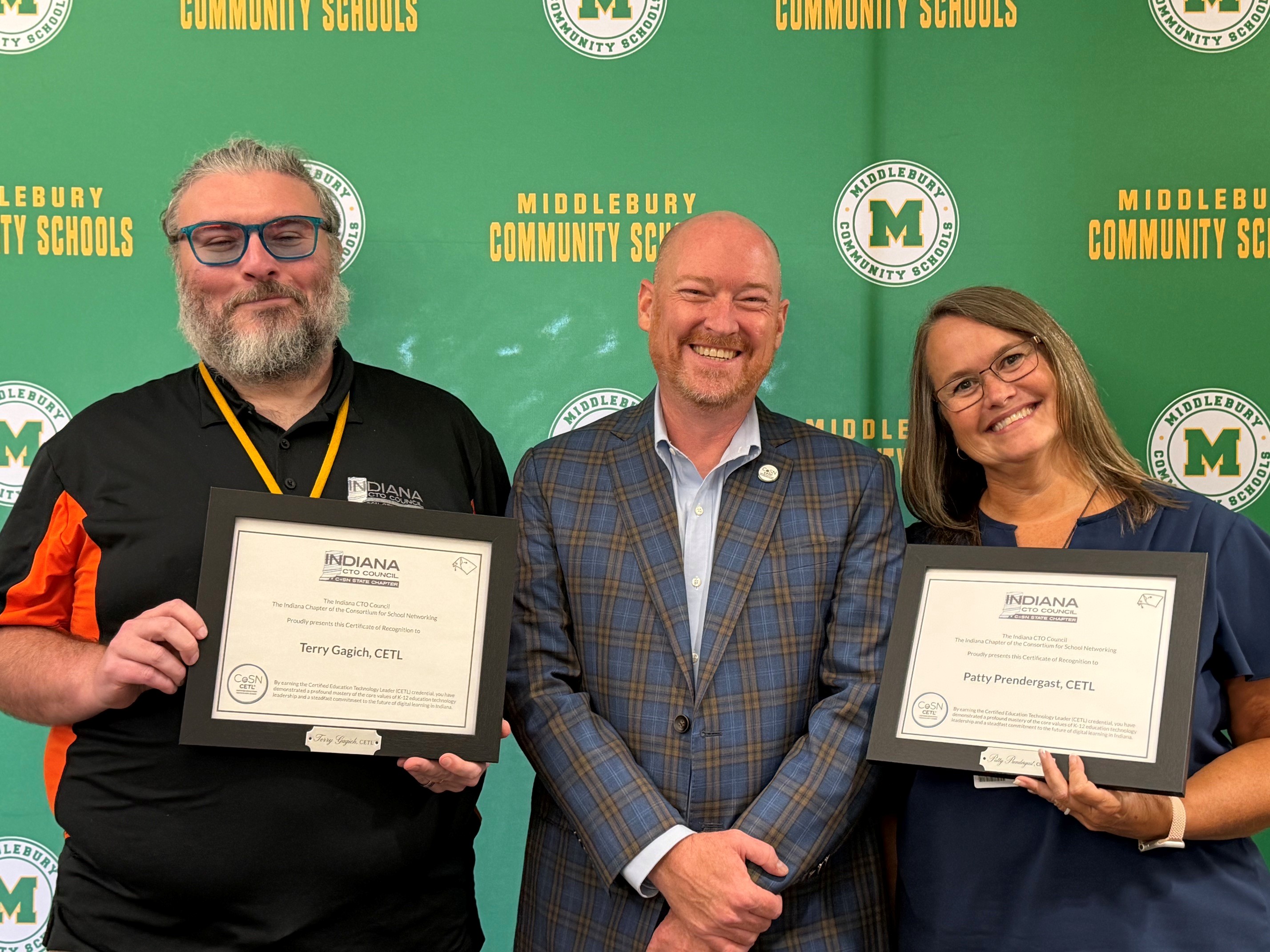 Three individuals smile as they hold framed certificates in front of a green banner with "MIDDLEBURY COMMUNITY SCHOOLS" text.