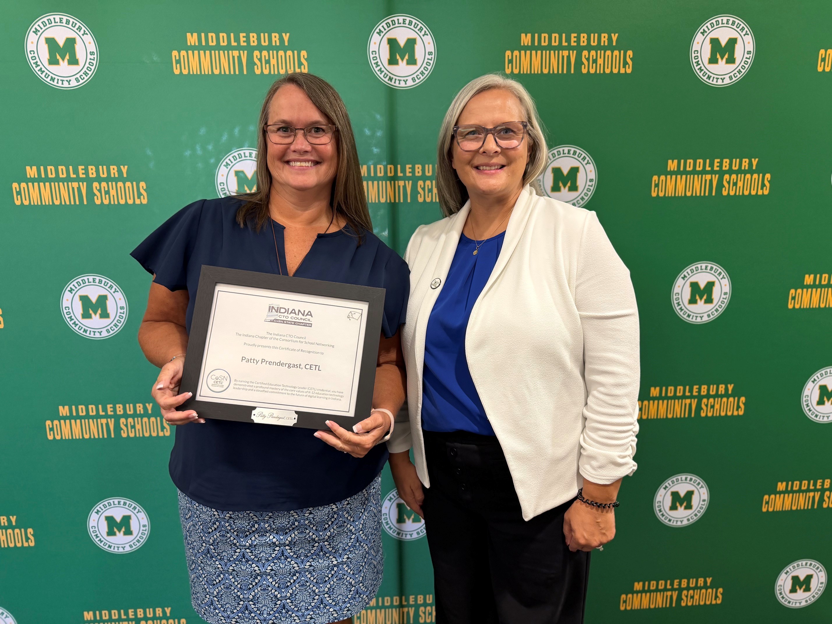 Two women stand in front of a green backdrop with "MIDDLEBURY COMMUNITY SCHOOLS" logos. The woman on the left holds a framed certificate.