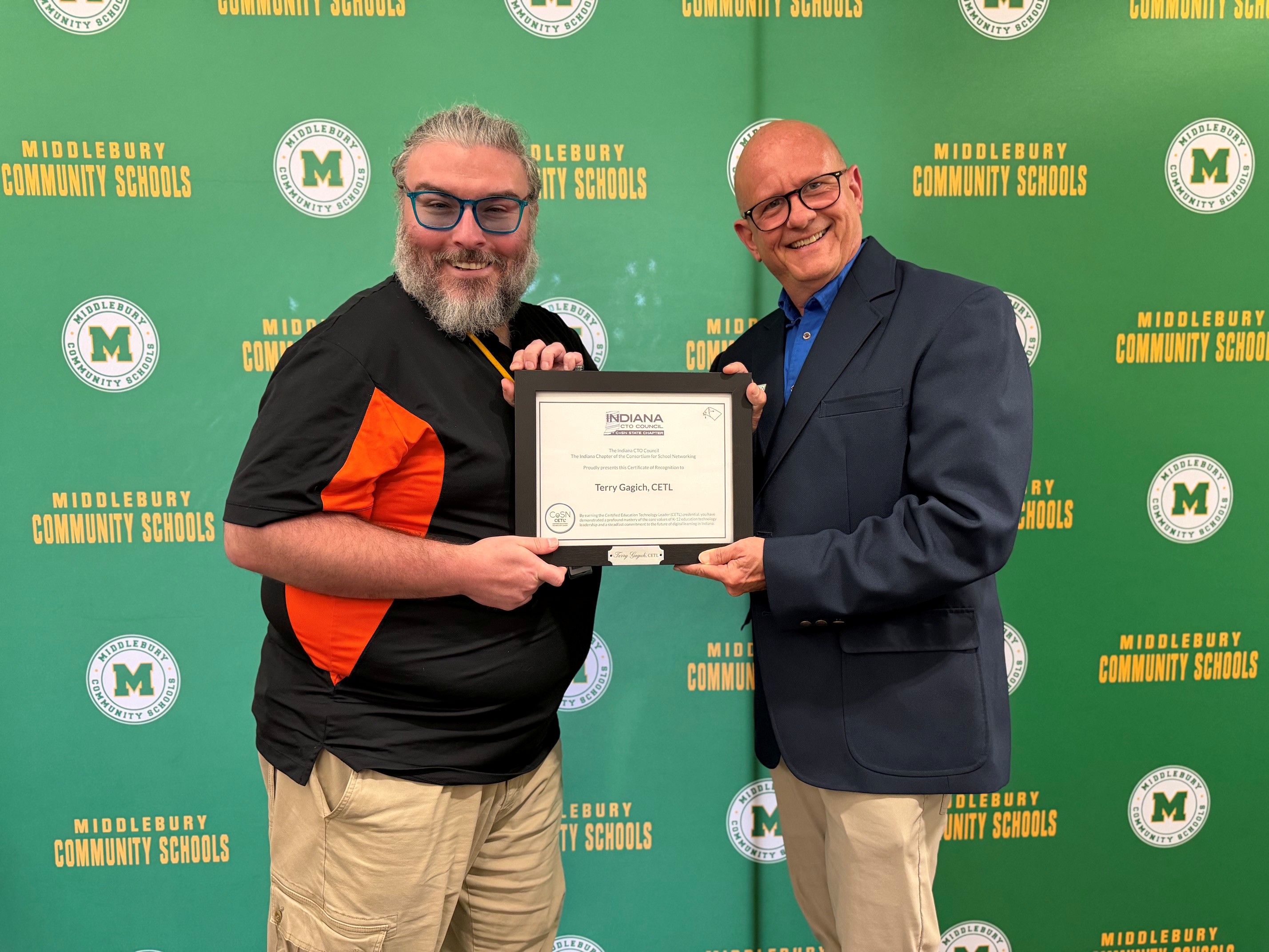 Two men smile as one presents a framed certificate to the other in front of a green backdrop with "MIDDLEBURY COMMUNITY SCHOOLS" logos.