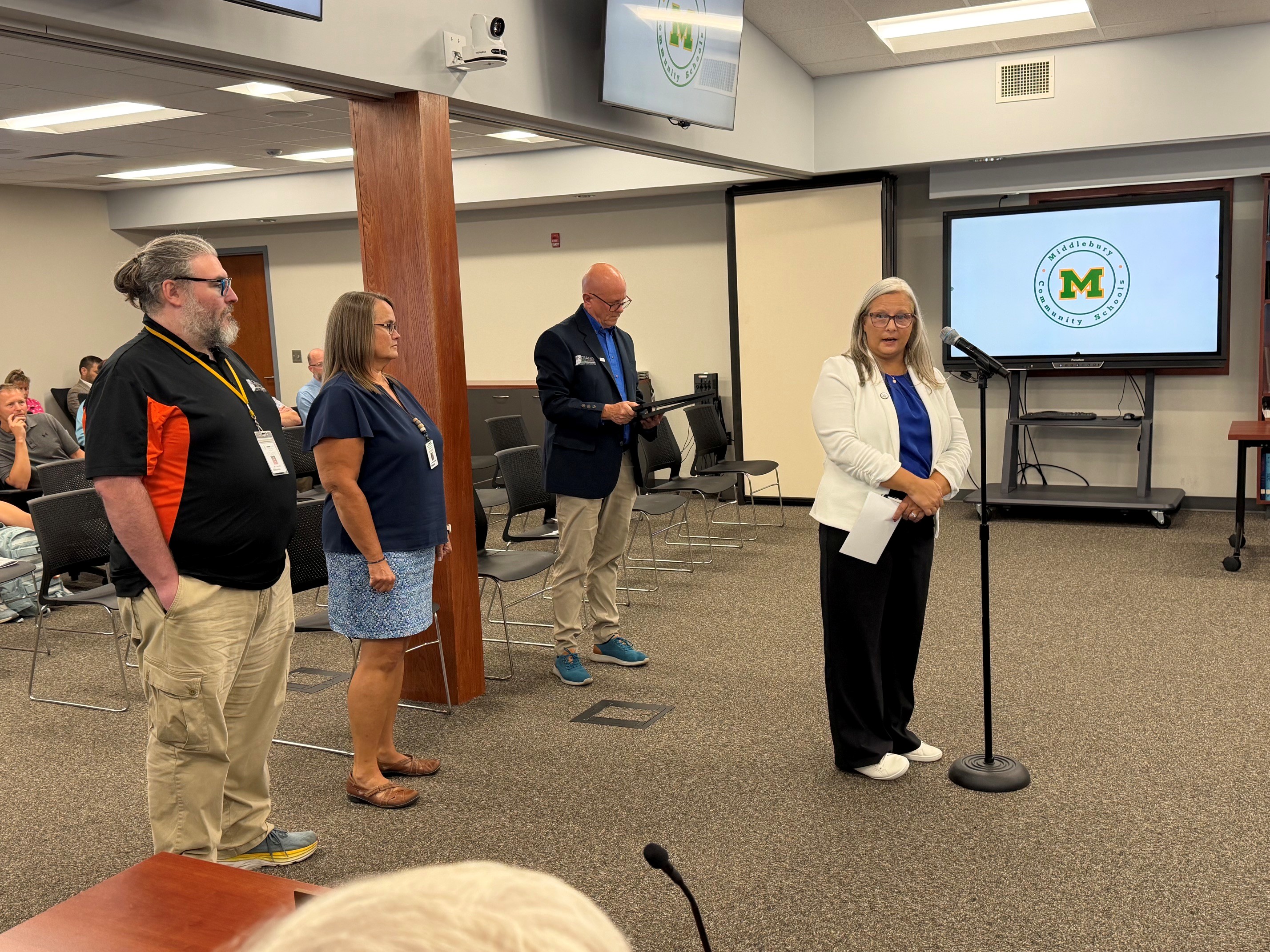 Four individuals stand in a meeting room, with a screen displaying the Middlebury Community Schools logo.