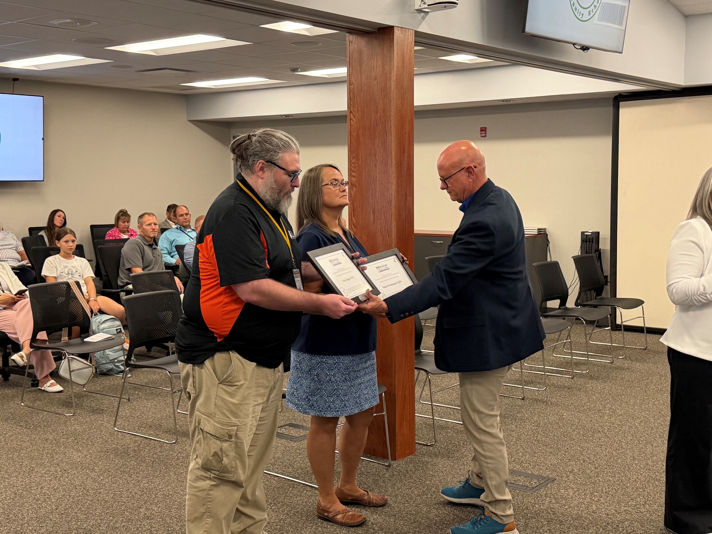 A man in a navy blazer presents a framed certificate to a woman in a blue skirt, while another man in an orange and black shirt looks on.