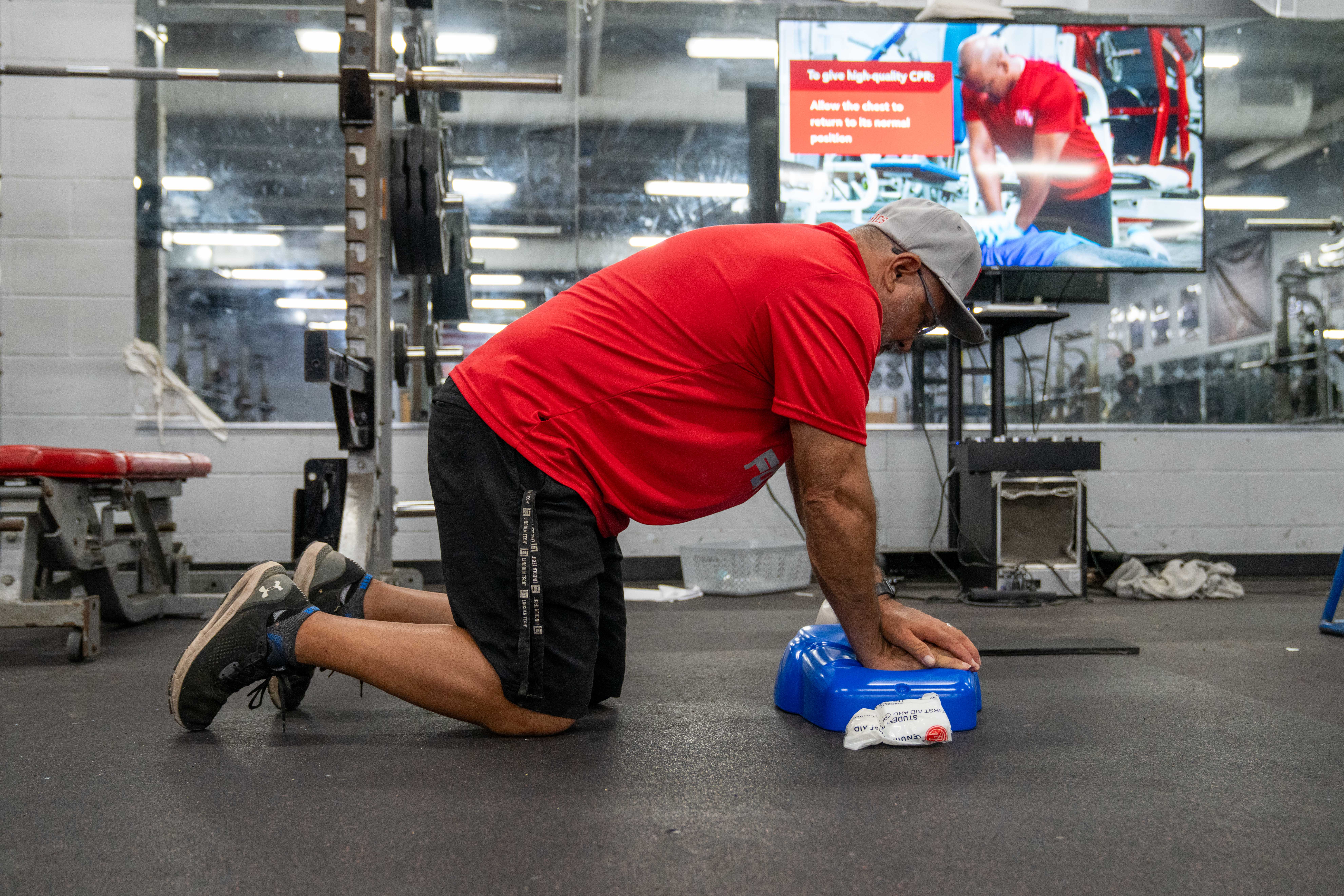A man in a red shirt and grey cap practices CPR on a blue training device in a gym.