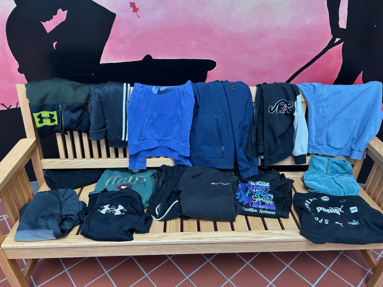 A wooden bench indoors holds a collection of folded and draped jackets and sweatshirts.