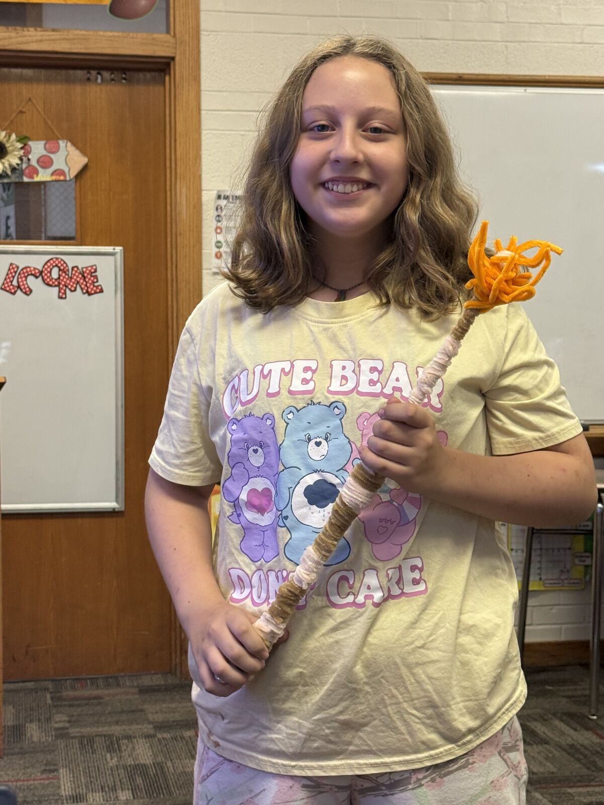 A smiling child holds up a long, handcrafted wand made with yarn and pipe cleaners in a classroom.