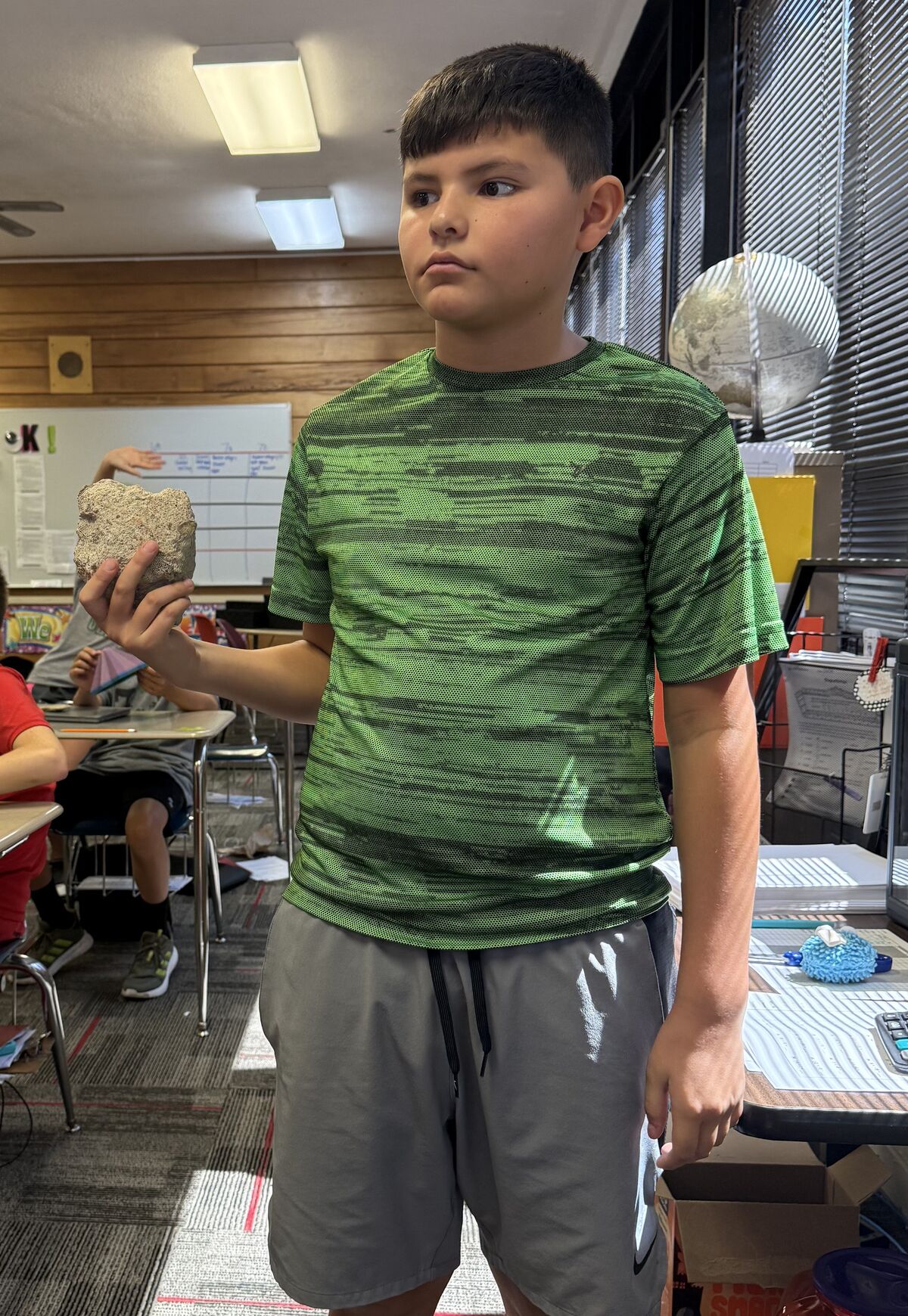 A boy in a green shirt and grey shorts stands in a classroom, holding a light-colored rock in his hand.