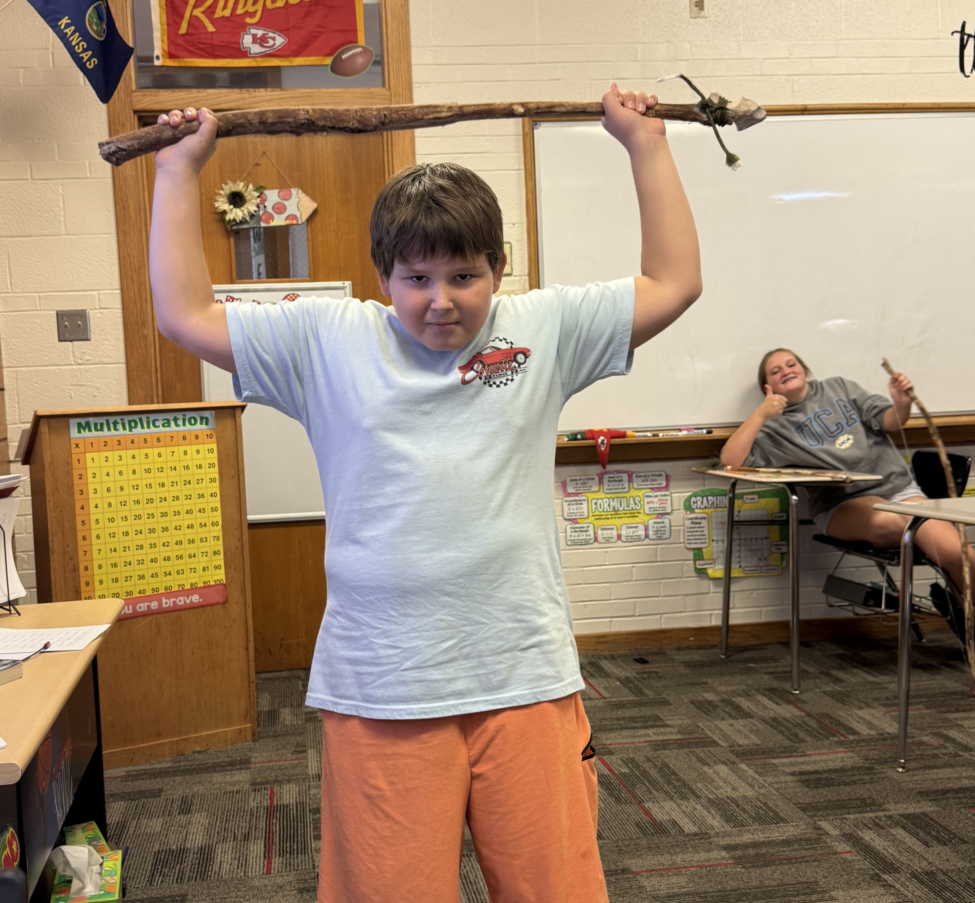 A young boy proudly holds a large wooden staff above his head in a brightly lit classroom.