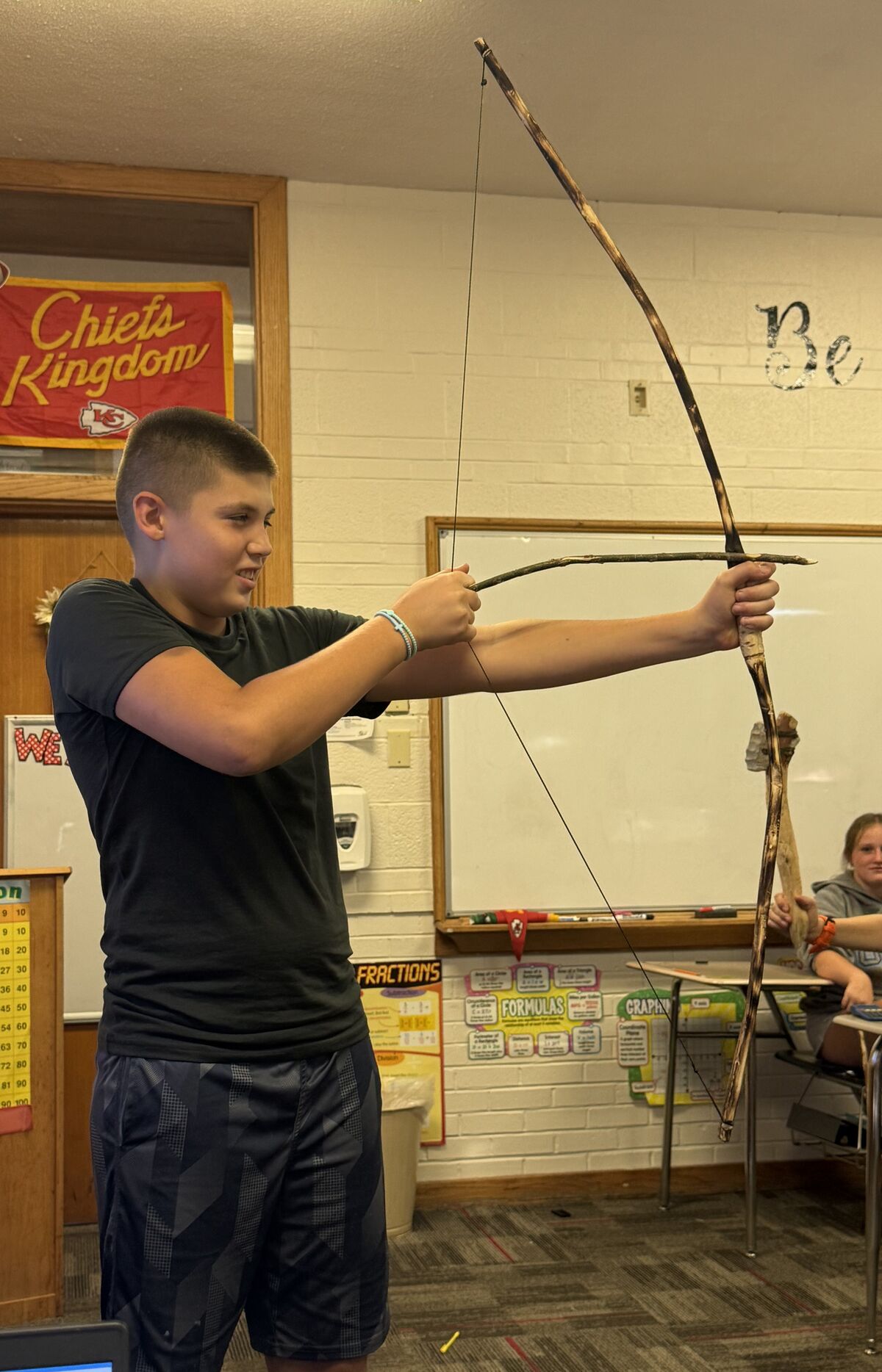 A young boy in a classroom holds a homemade wooden bow and arrow, preparing to draw it.