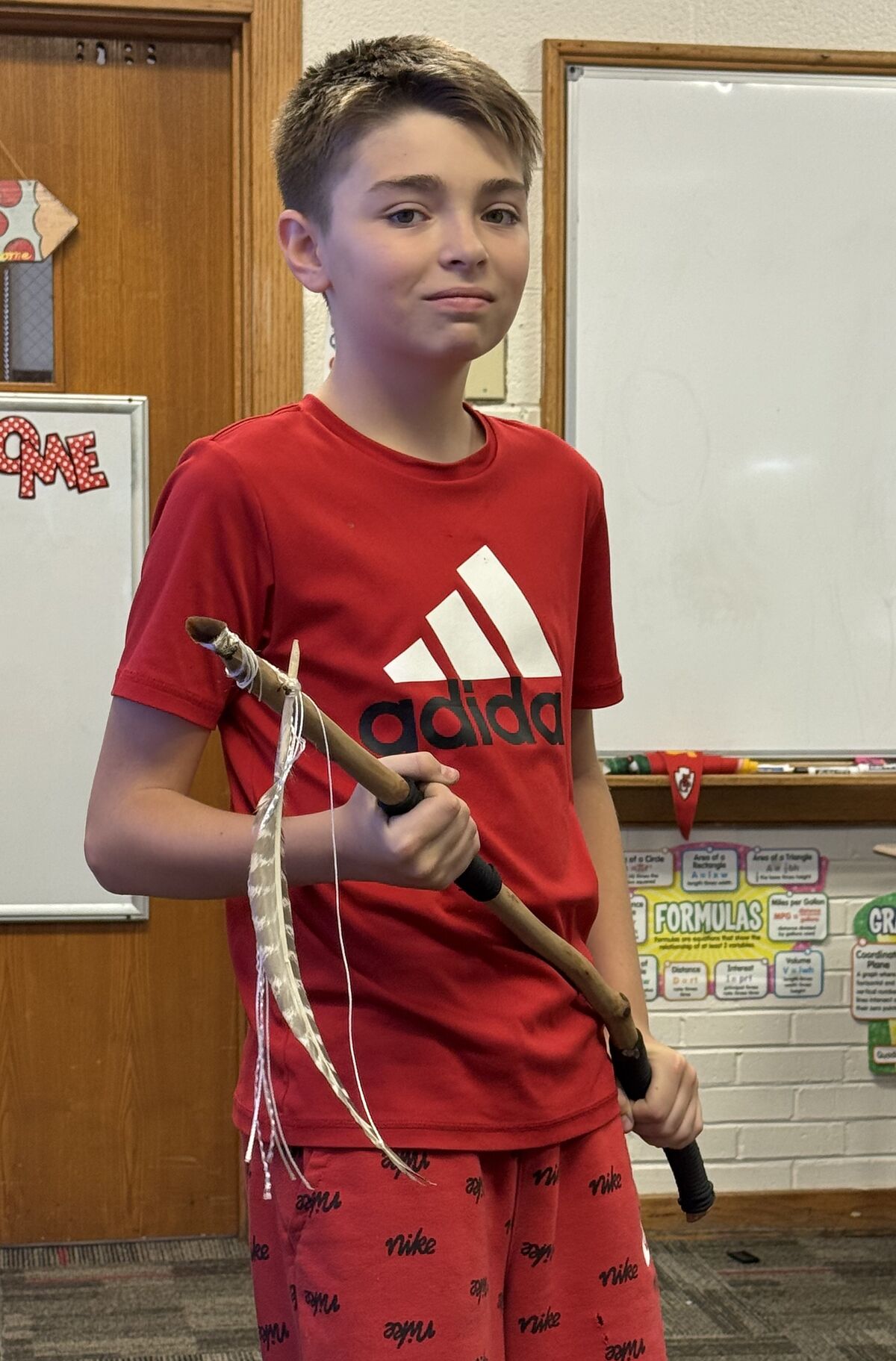 A young boy in a red Adidas t-shirt holds a decorated stick inside a classroom.