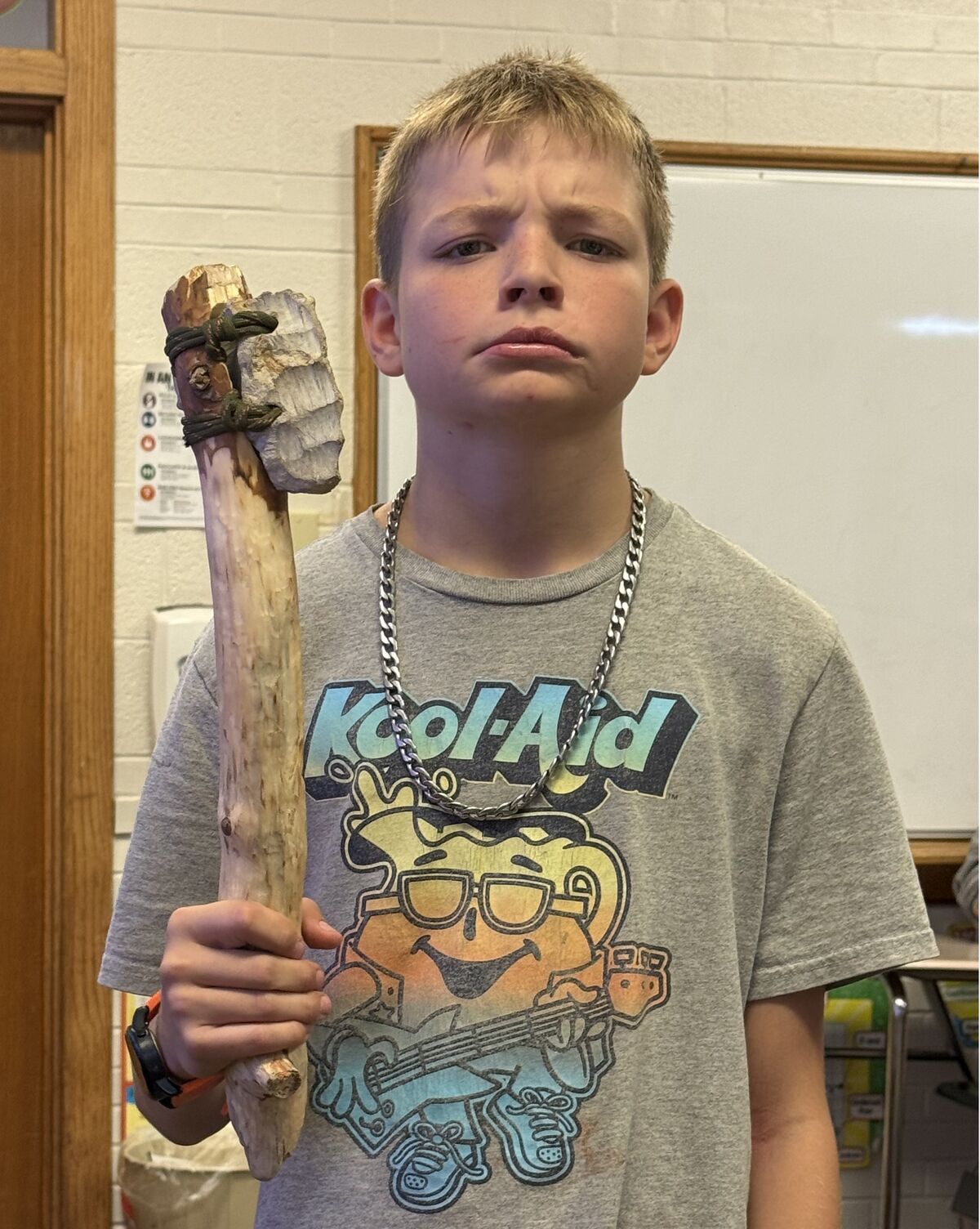 A young person in a grey Kool-Aid graphic t-shirt holds a homemade stone axe in an indoor setting.