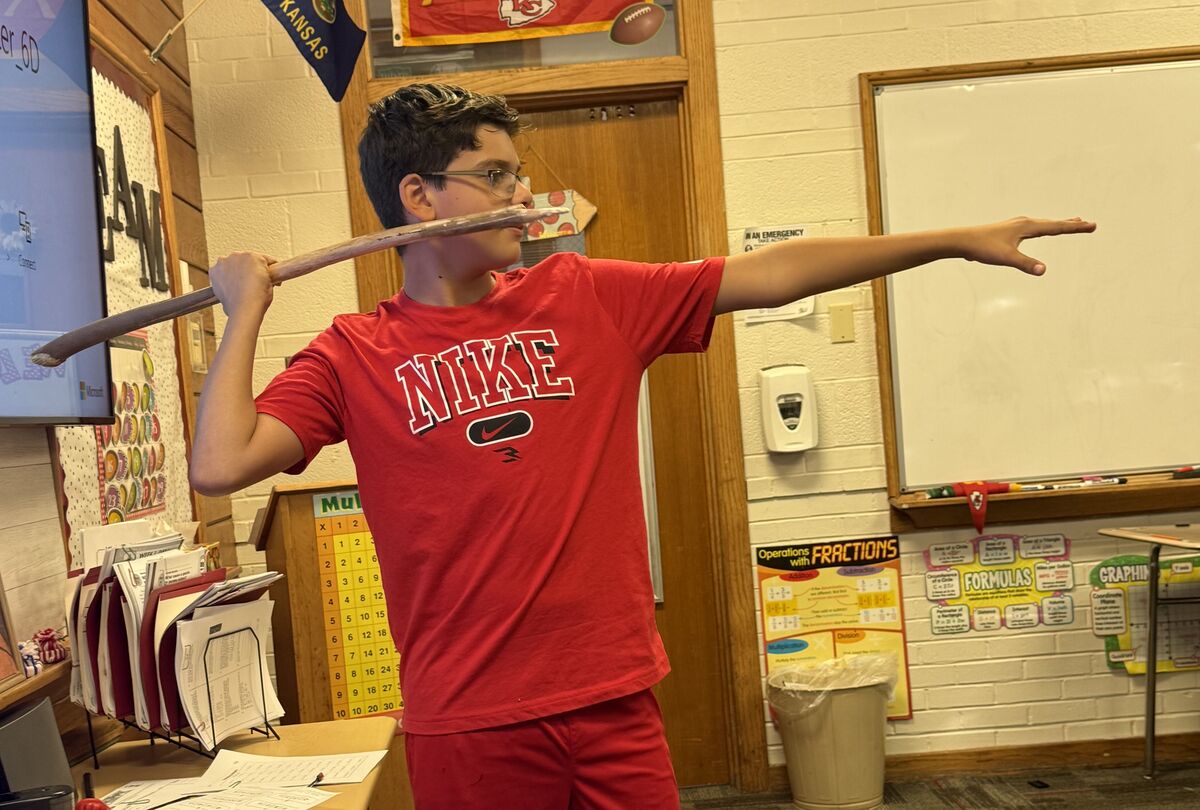 A student in a red Nike shirt poses in a classroom, pretending to shoot a bow and arrow with a wooden stick.