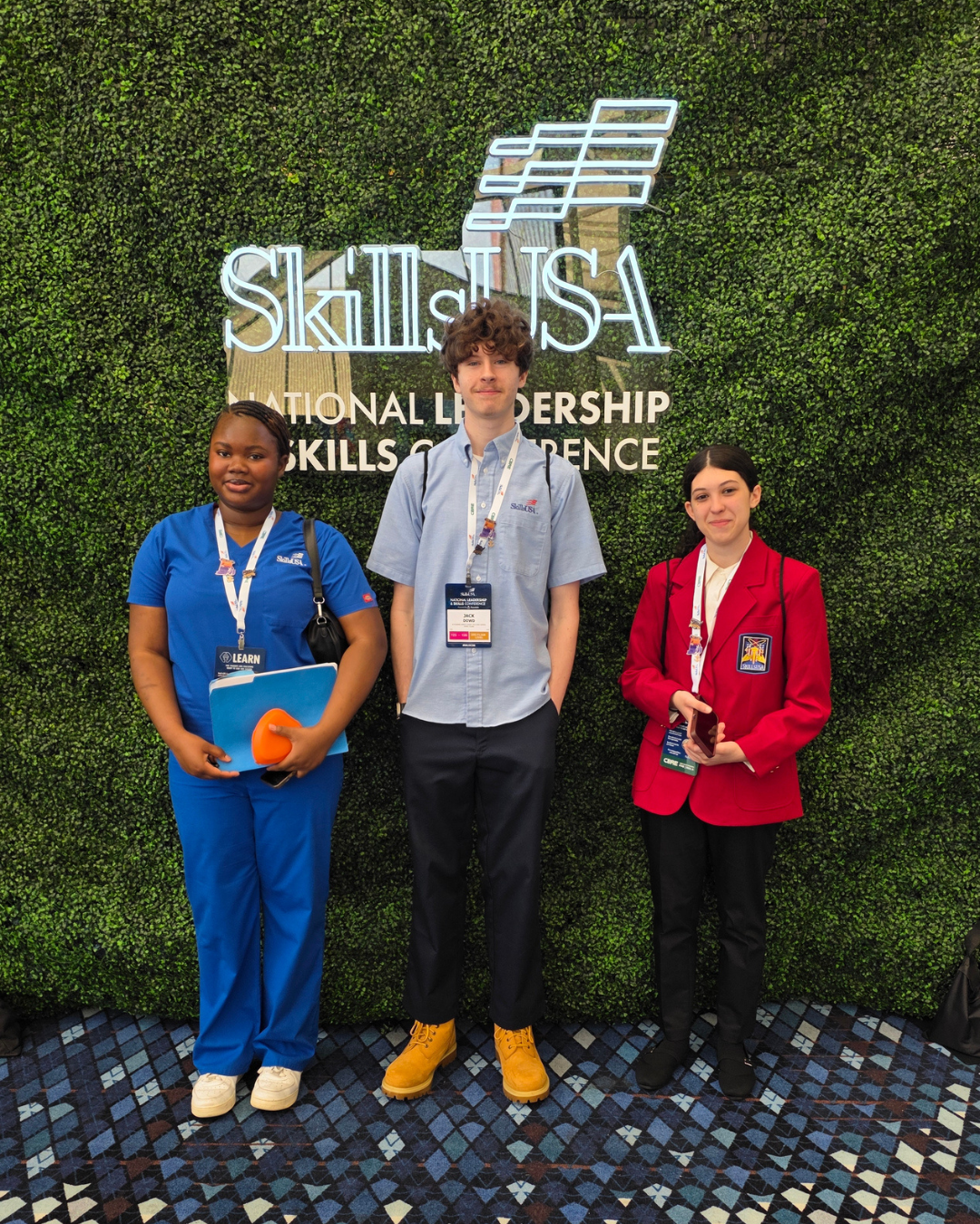Three young people stand in front of a green wall with a neon "SkillsUSA" logo.