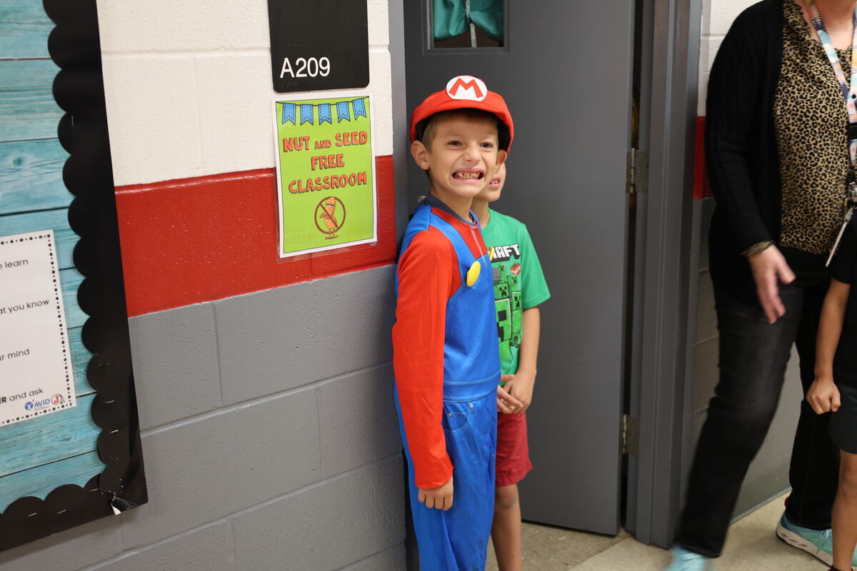 A smiling child wearing a Super Mario costume stands near a classroom door in a school hallway.
