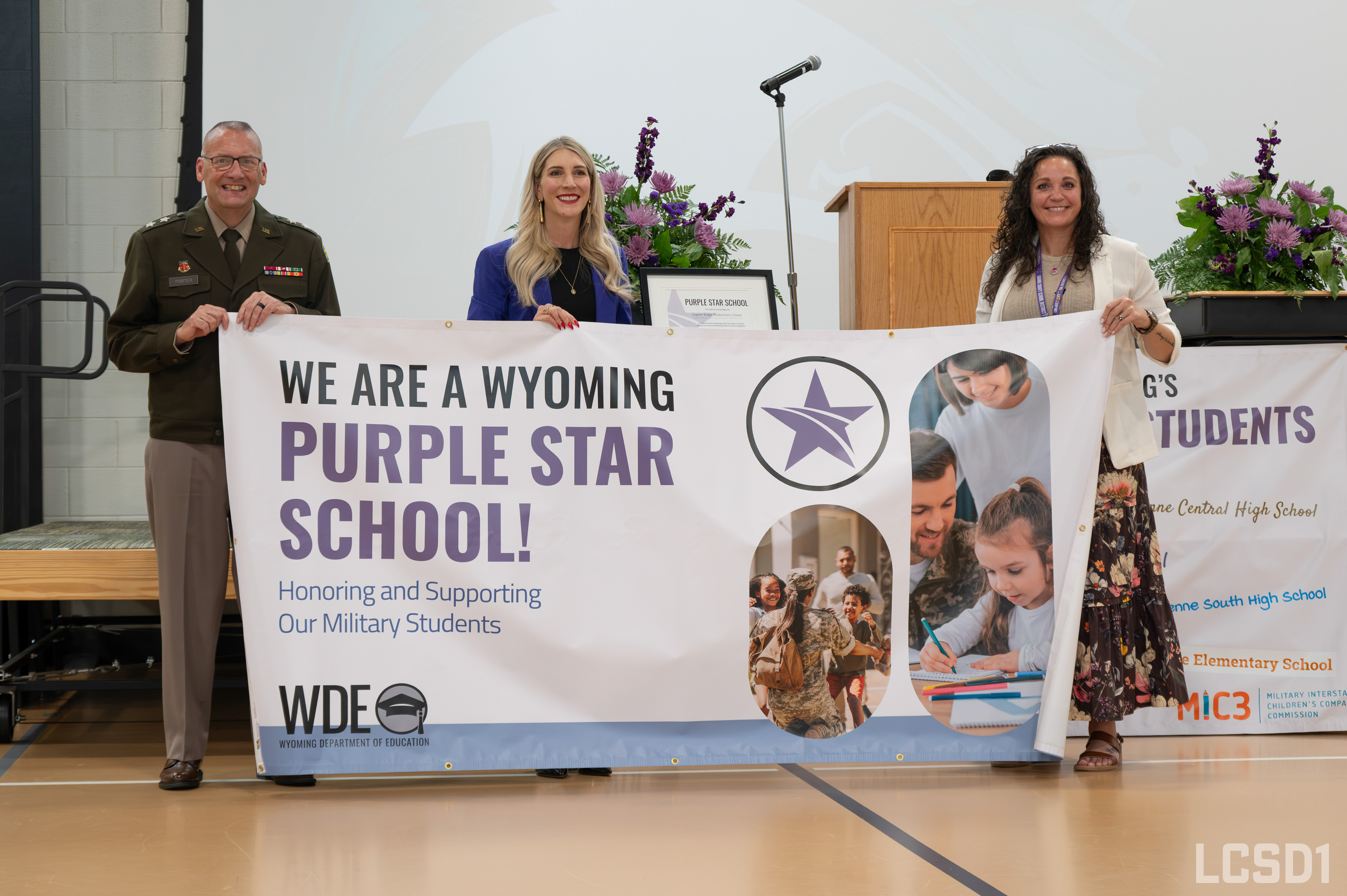 Three people proudly hold a large banner that reads "We Are a Wyoming Purple Star School! Honoring and Supporting Our Military Students."