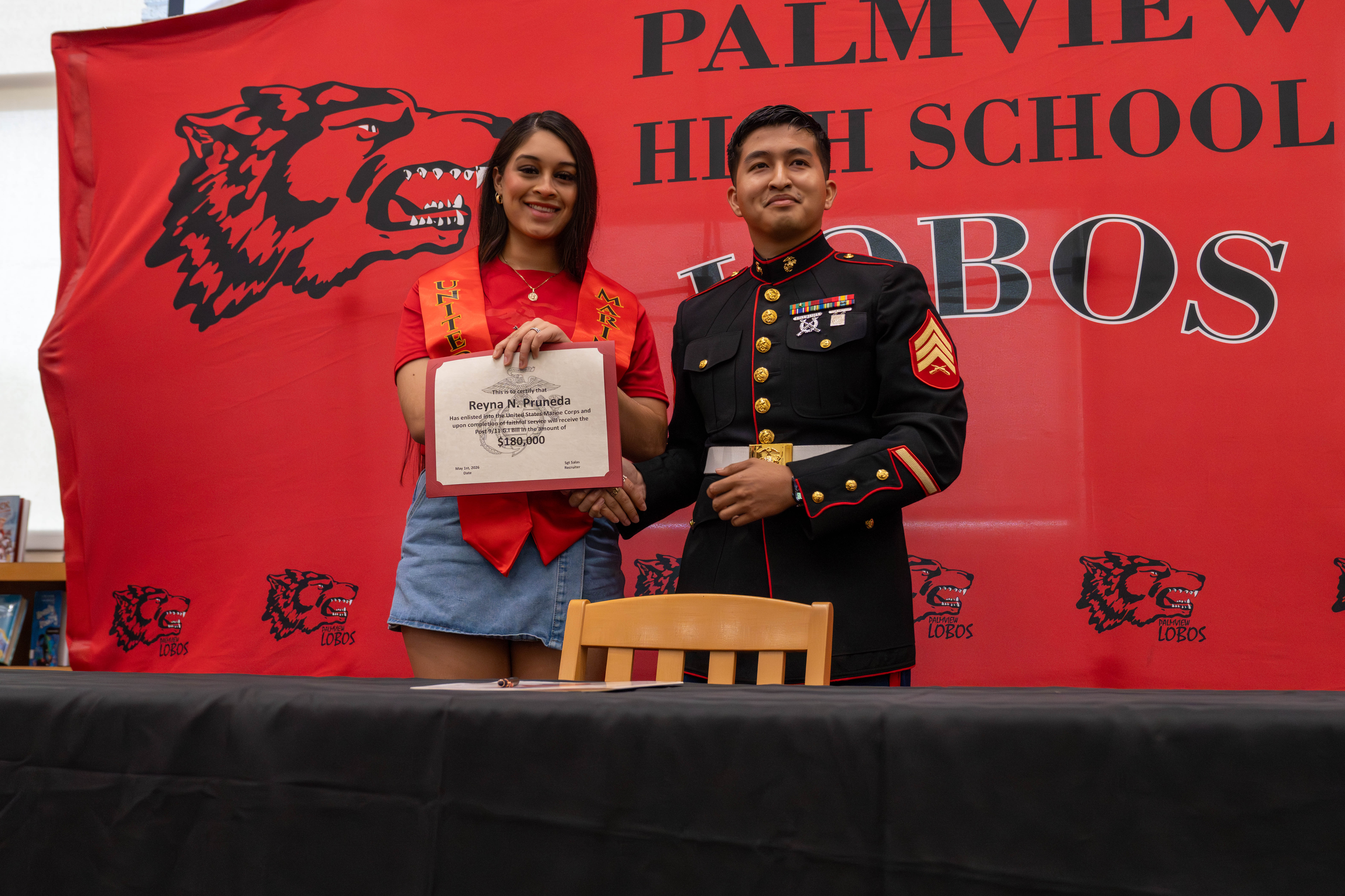 A young woman in a red shirt and sash receives a certificate from a Marine in uniform.