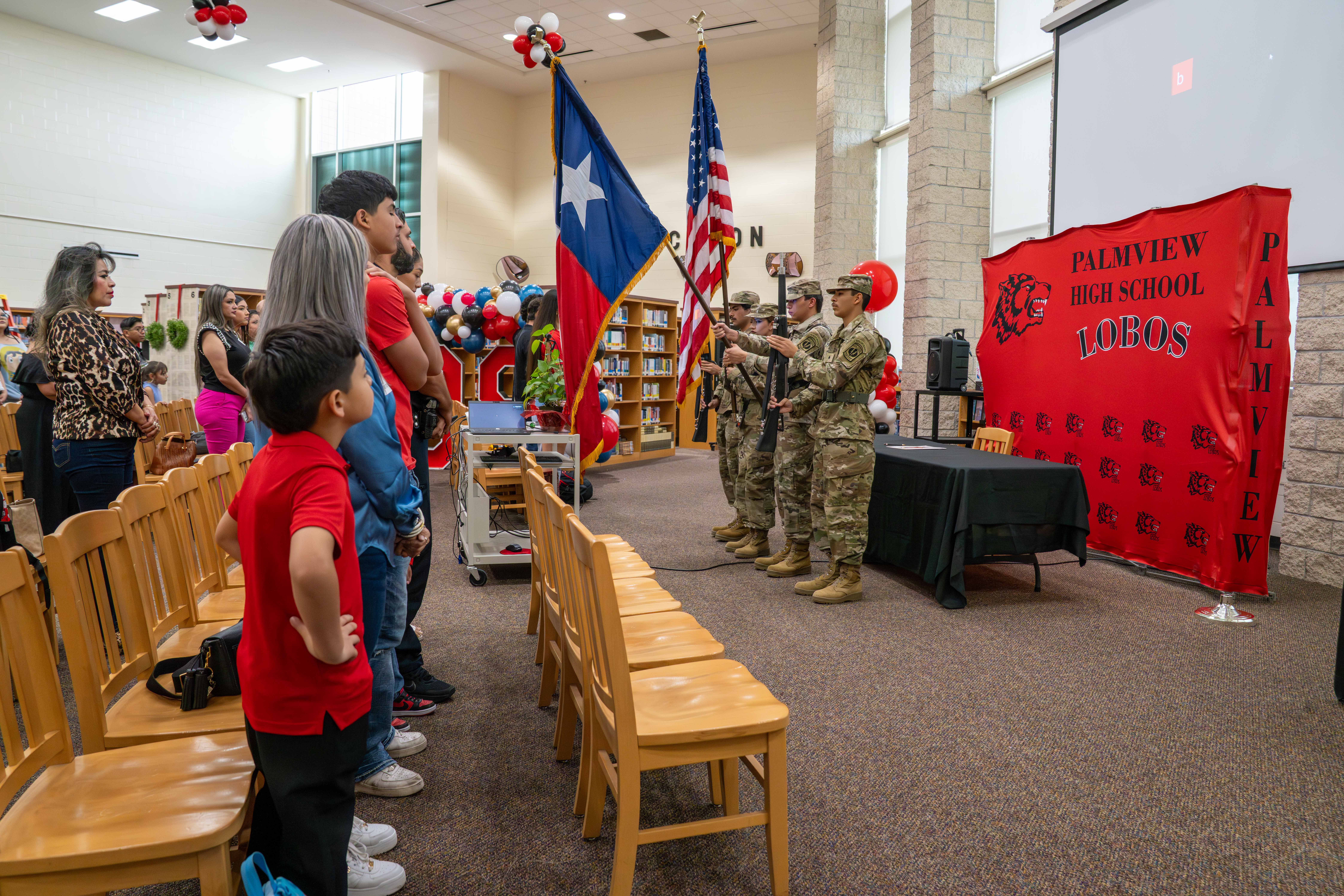 A group of students and adults stand in an auditorium, facing a color guard presenting flags.