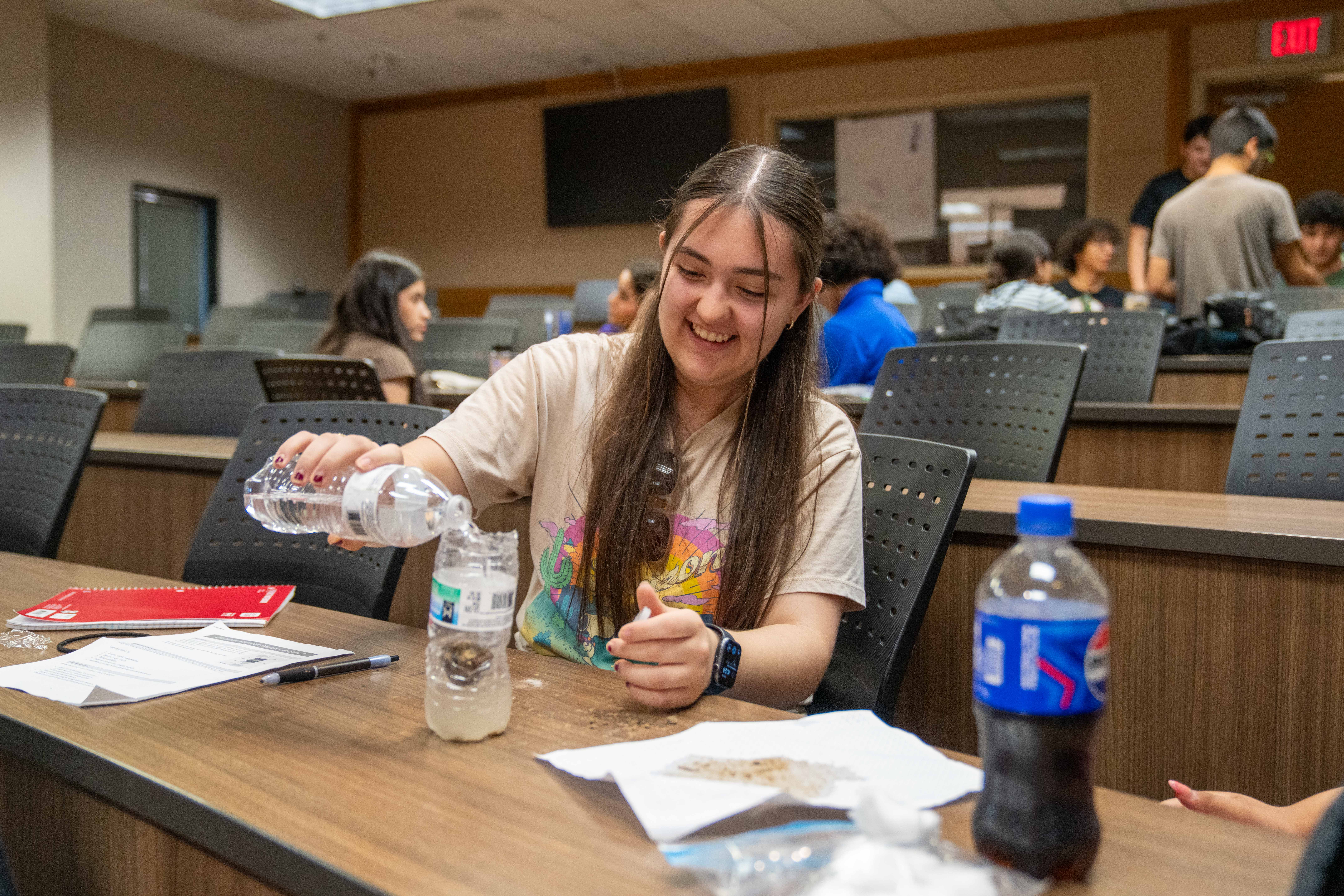 A young woman smiles as she pours water from a bottle into another bottle on a classroom desk.