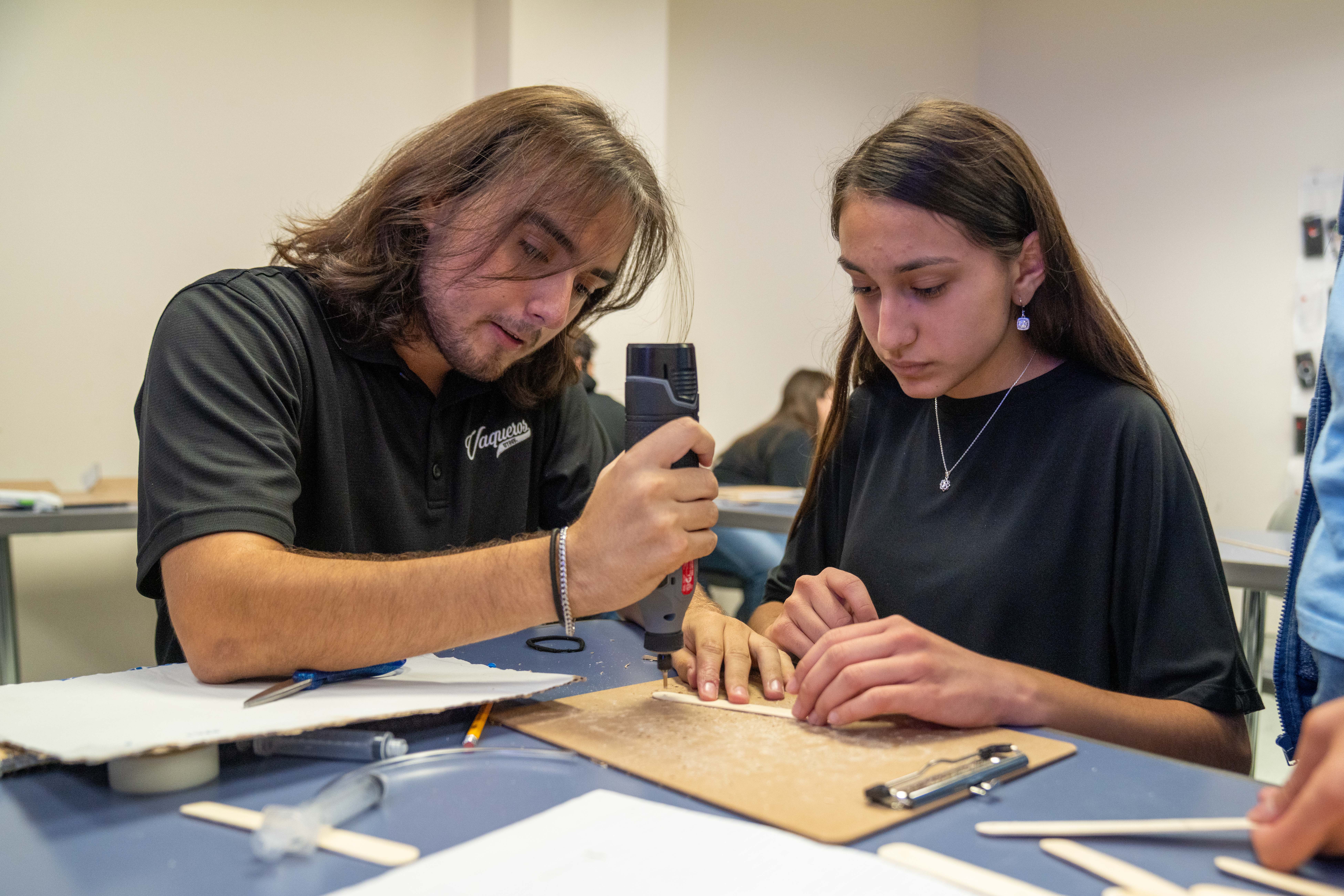 Two students collaborate on a craft project, one using a rotary tool while the other holds a wooden stick.