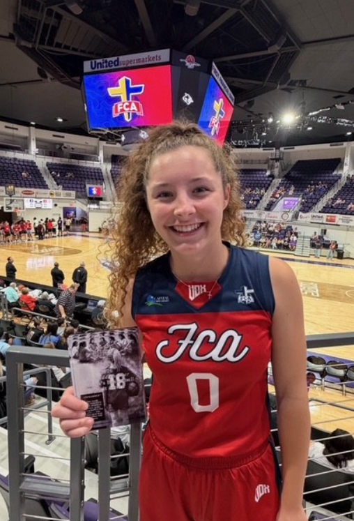 A young woman in a red basketball uniform smiles, holding a card with a black and white photo.