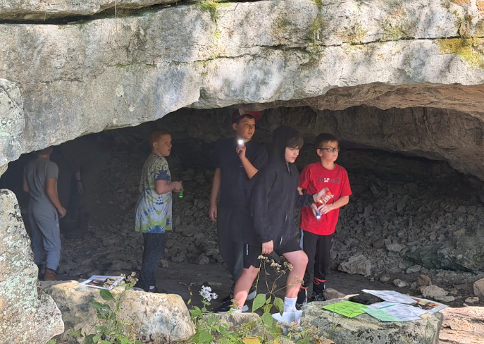 A group of children explore the entrance of a rocky cave, holding flashlights.
