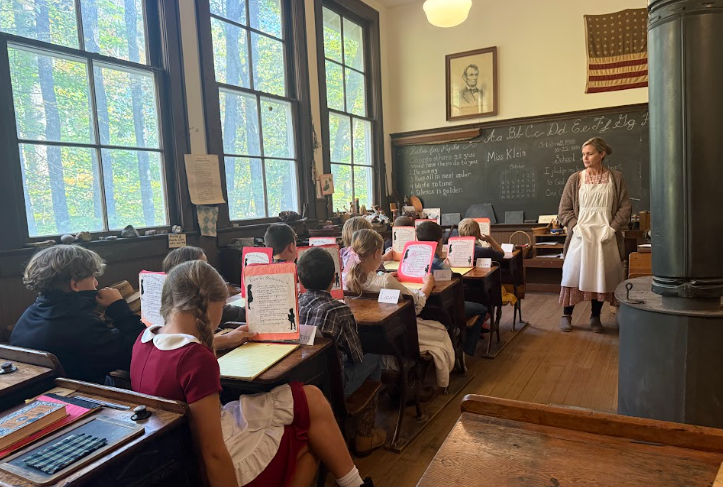 A teacher stands in front of a historical one-room schoolhouse while children sit at wooden desks reading.