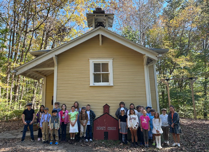 A large group of children poses in front of the yellow Honey Creek schoolhouse surrounded by autumn trees.