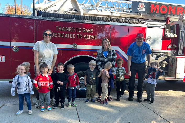 A group of children and adults pose in front of a large red fire truck reading "TRAINED TO SAVE, DEDICATED TO SERVE."
