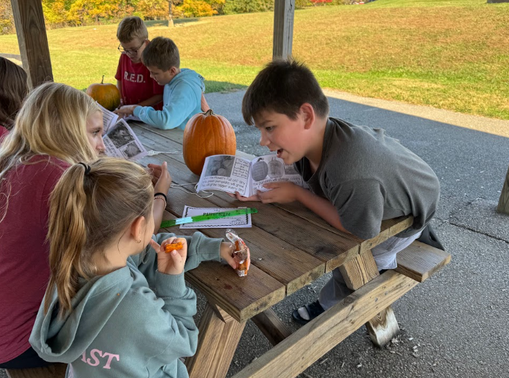 A group of children sit around a wooden picnic table outdoors, reading small booklets and interacting with each other.