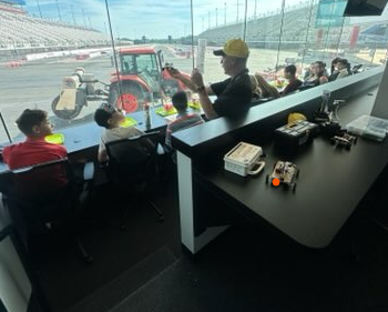 A group of people, including children and adults, sit at a table in a room overlooking a race track with a tractor in the distance.