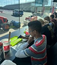 A young person with glasses works on a craft project at a table, with a race track visible through a large window.