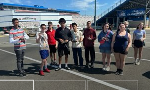 A group of ten young people stand together outdoors in front of a grandstand at a race track.