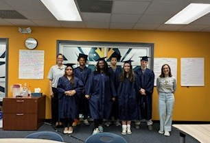 A group of graduates in navy blue gowns and caps stand together, smiling.