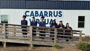 Six graduates in navy blue caps and gowns stand on a wooden ramp in front of a building with "CABARRUS VIRTUAL" signage.