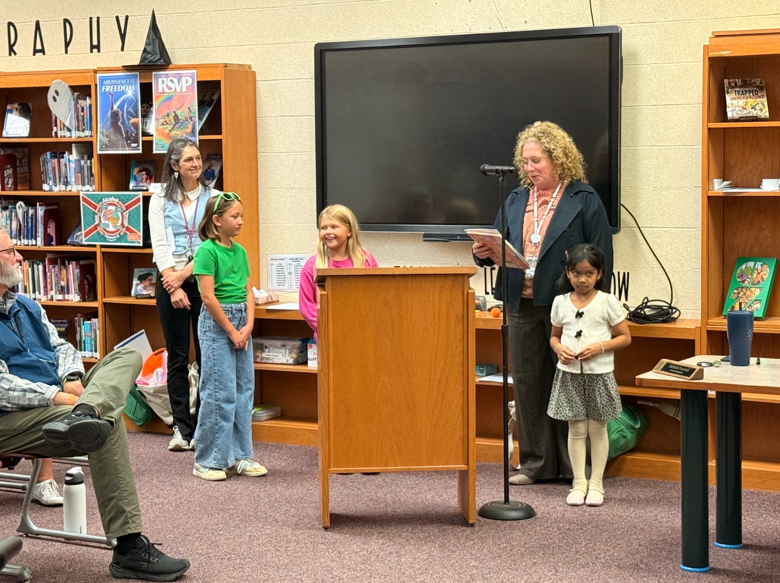 A group of people gathered in a library setting, with a speaker at a wooden podium reading a book into a microphone.