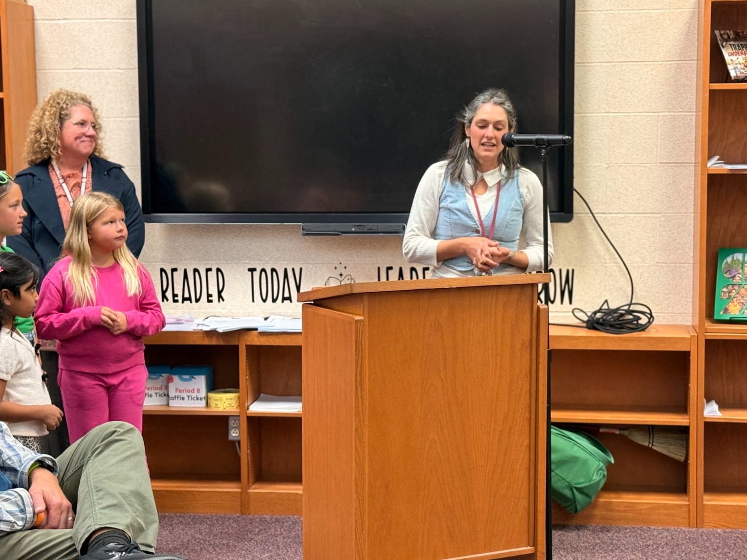 A woman speaks at a wooden podium in a library, while children and adults watch from the audience.