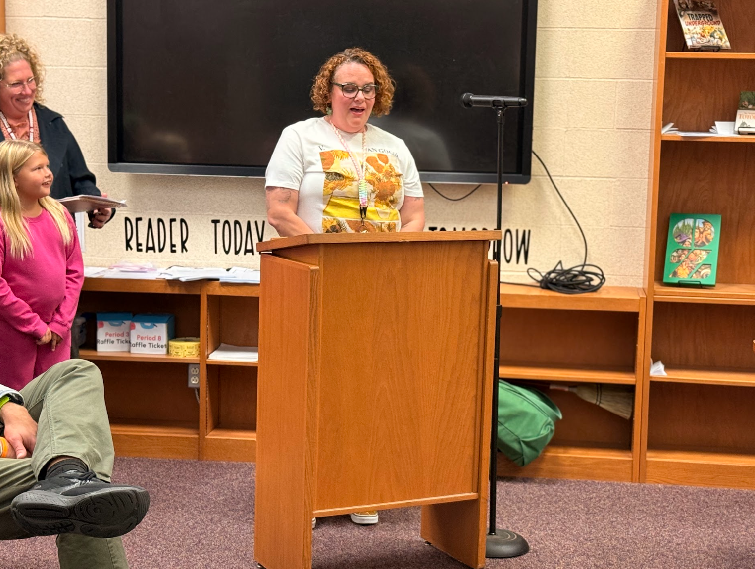 A woman speaks at a wooden podium in a library, with a microphone in front of her.