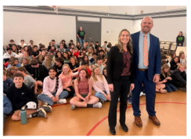 A male and female adult stand in front of a large group of seated students in a gymnasium.
