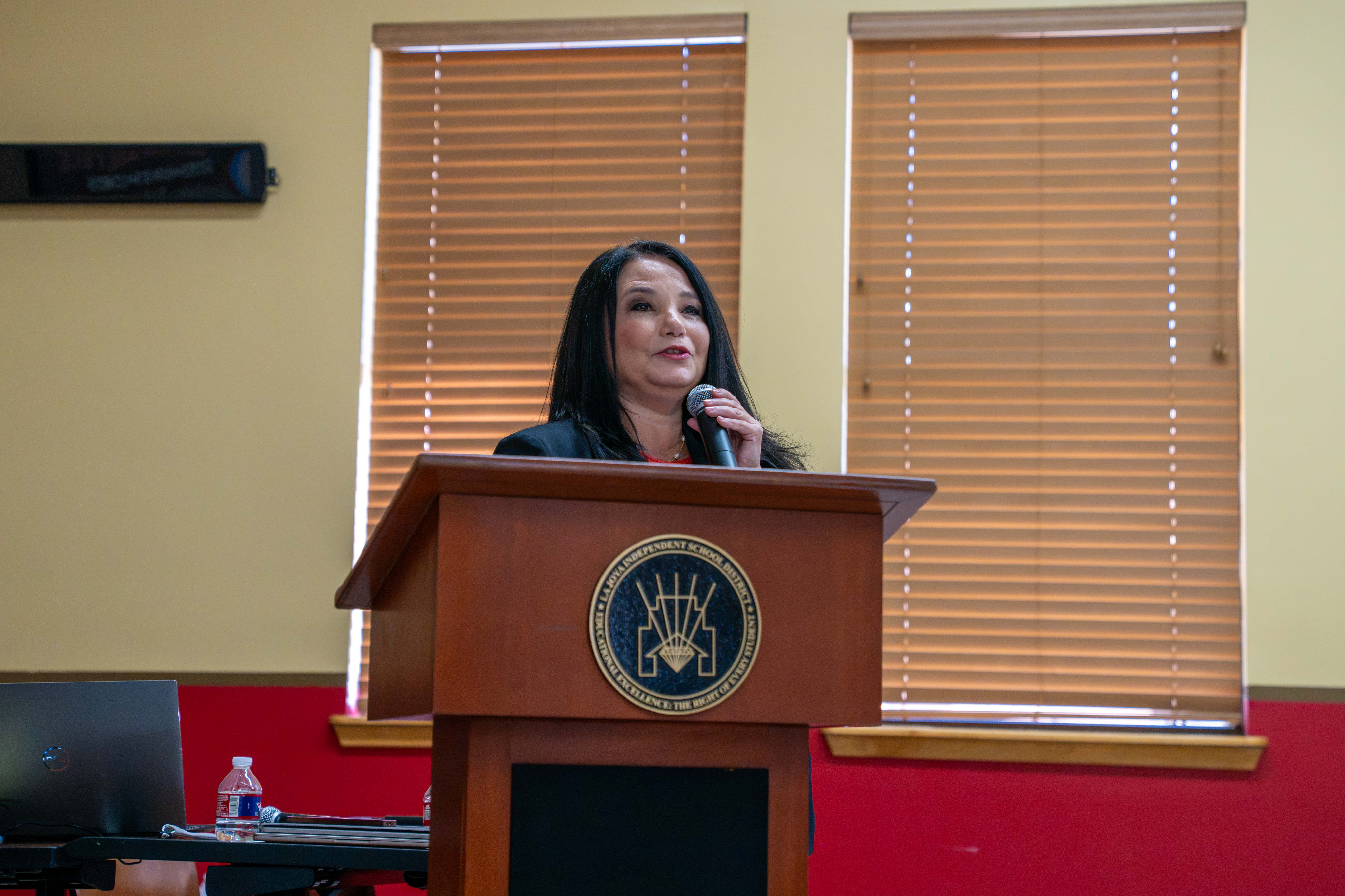 A woman stands at a wooden podium speaking into a microphone in a room with two windows with closed blinds.