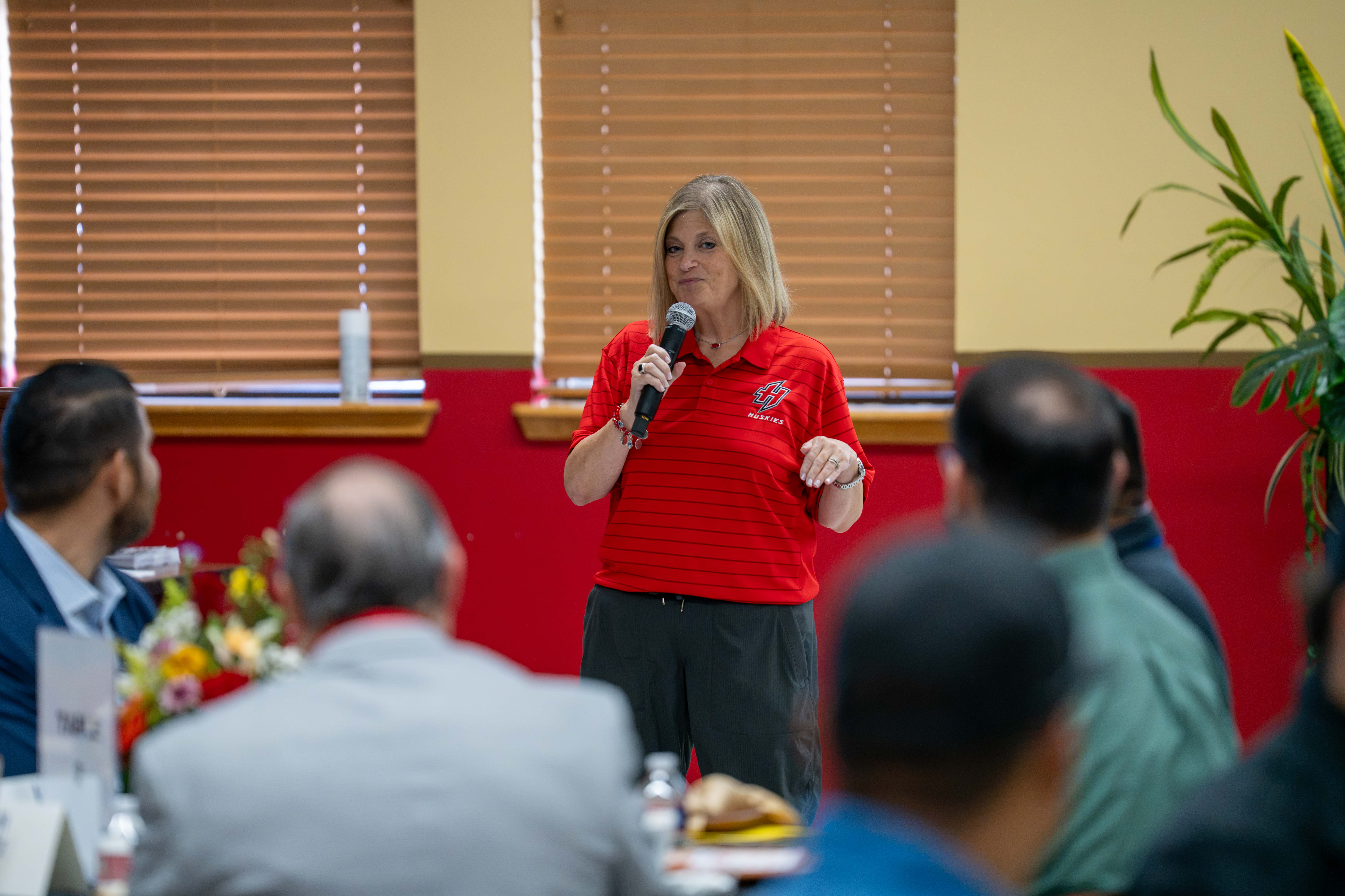 A woman wearing a red polo shirt speaks into a microphone before an attentive seated audience.