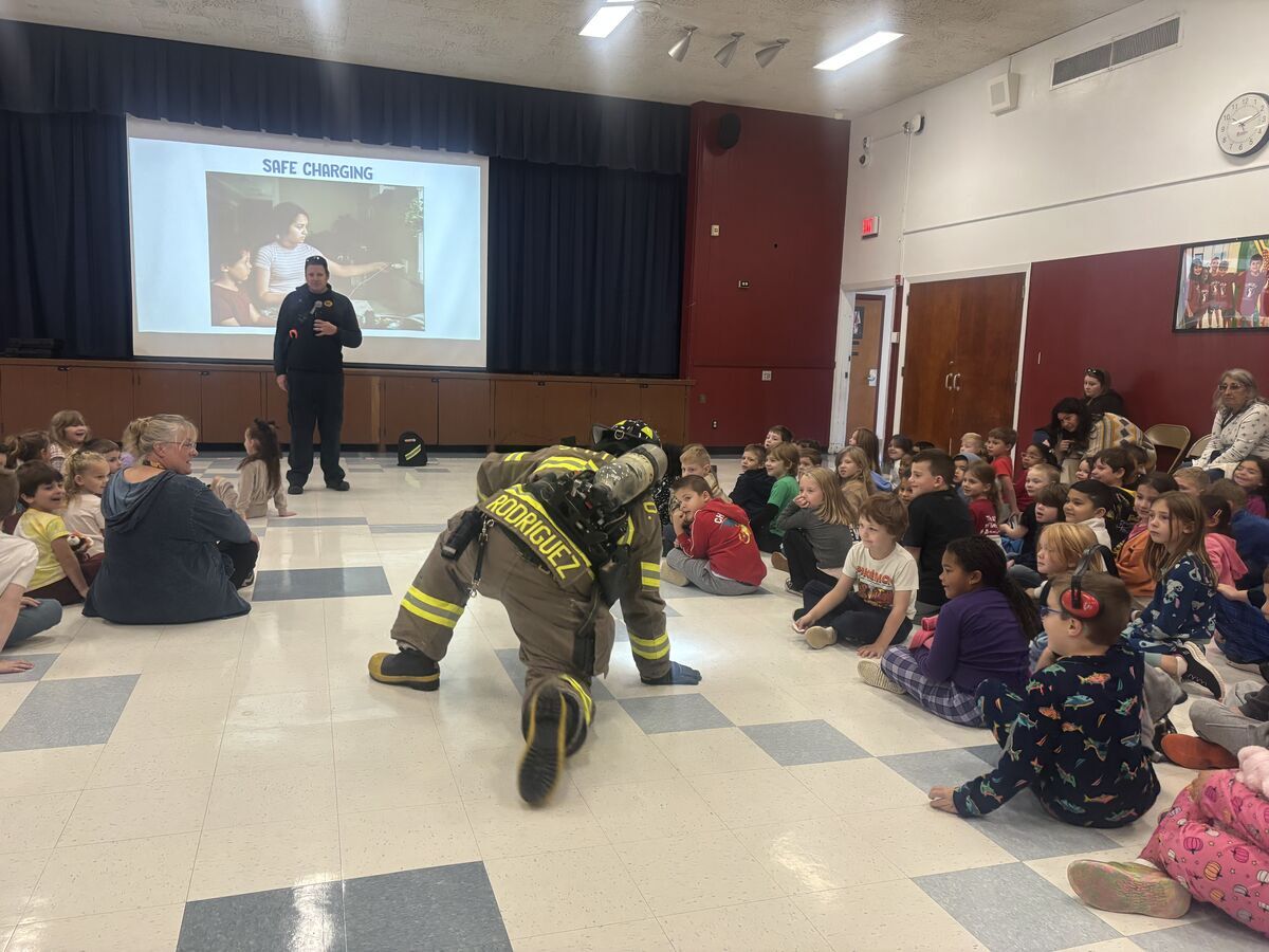 A firefighter in full gear kneels before a large group of attentive school children seated on a school auditorium floor.