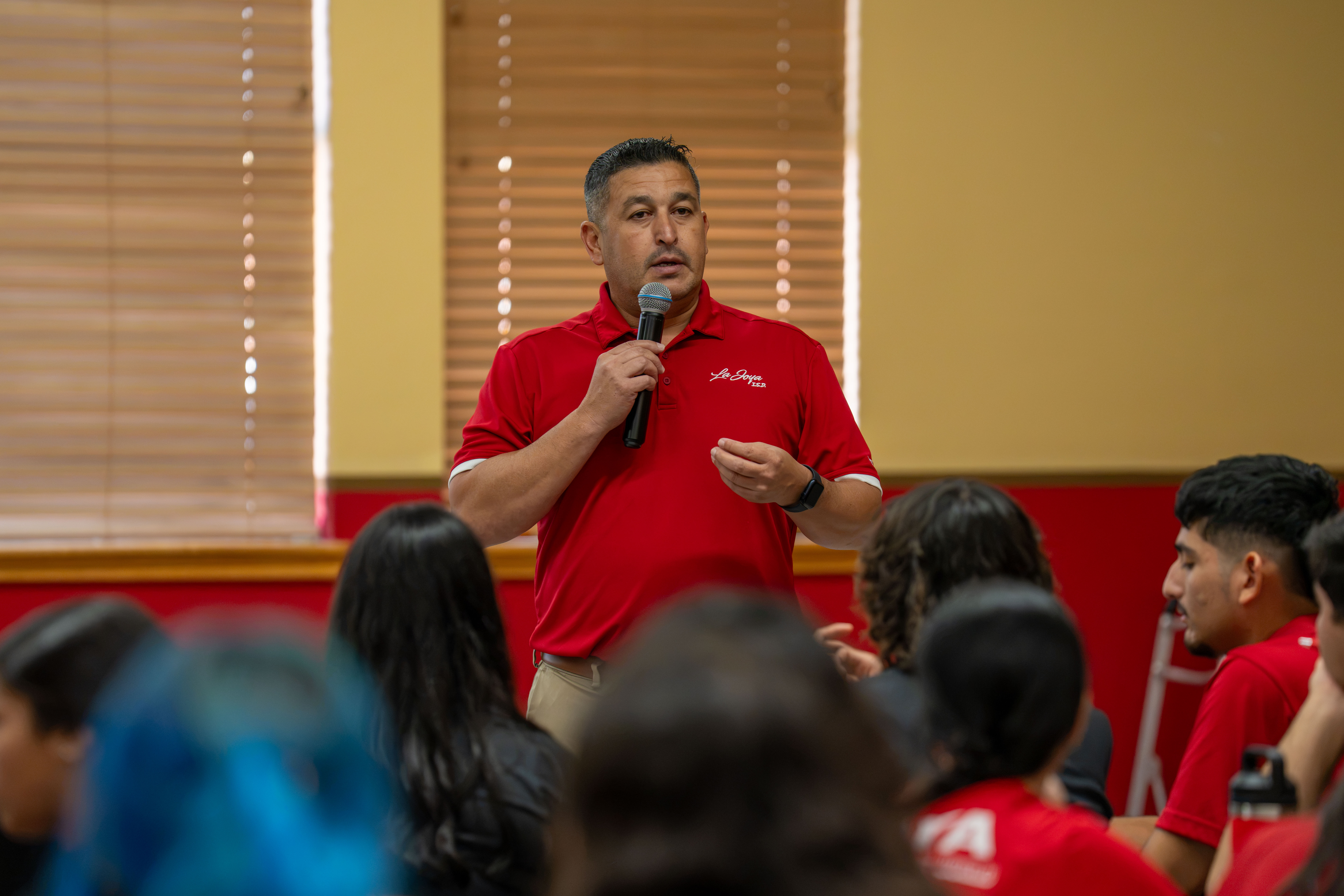 A man in a red polo shirt speaks into a microphone while addressing a seated audience indoors.