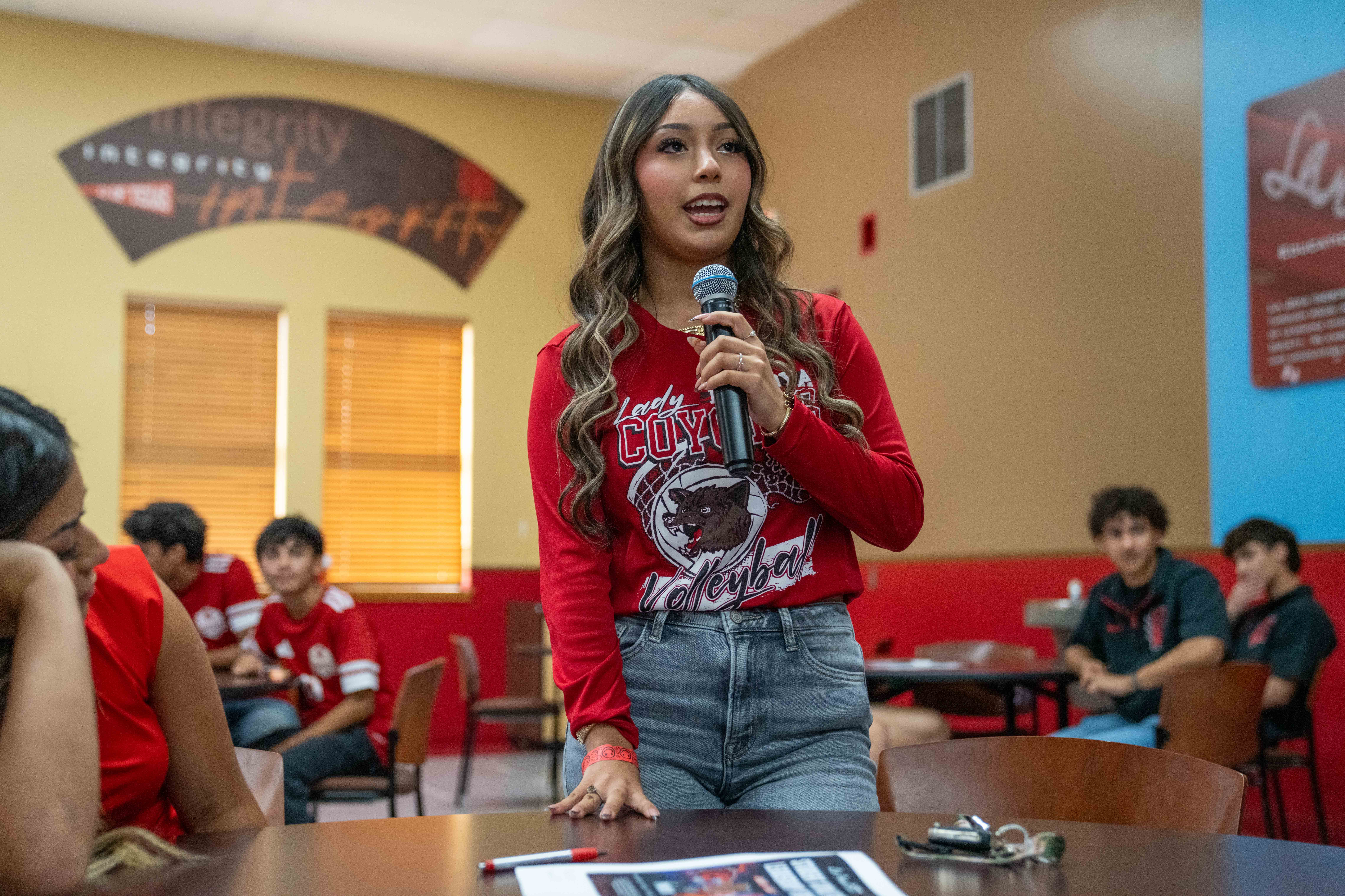 A young woman in a red long-sleeve shirt stands speaking into a microphone in a room with other people seated at tables.