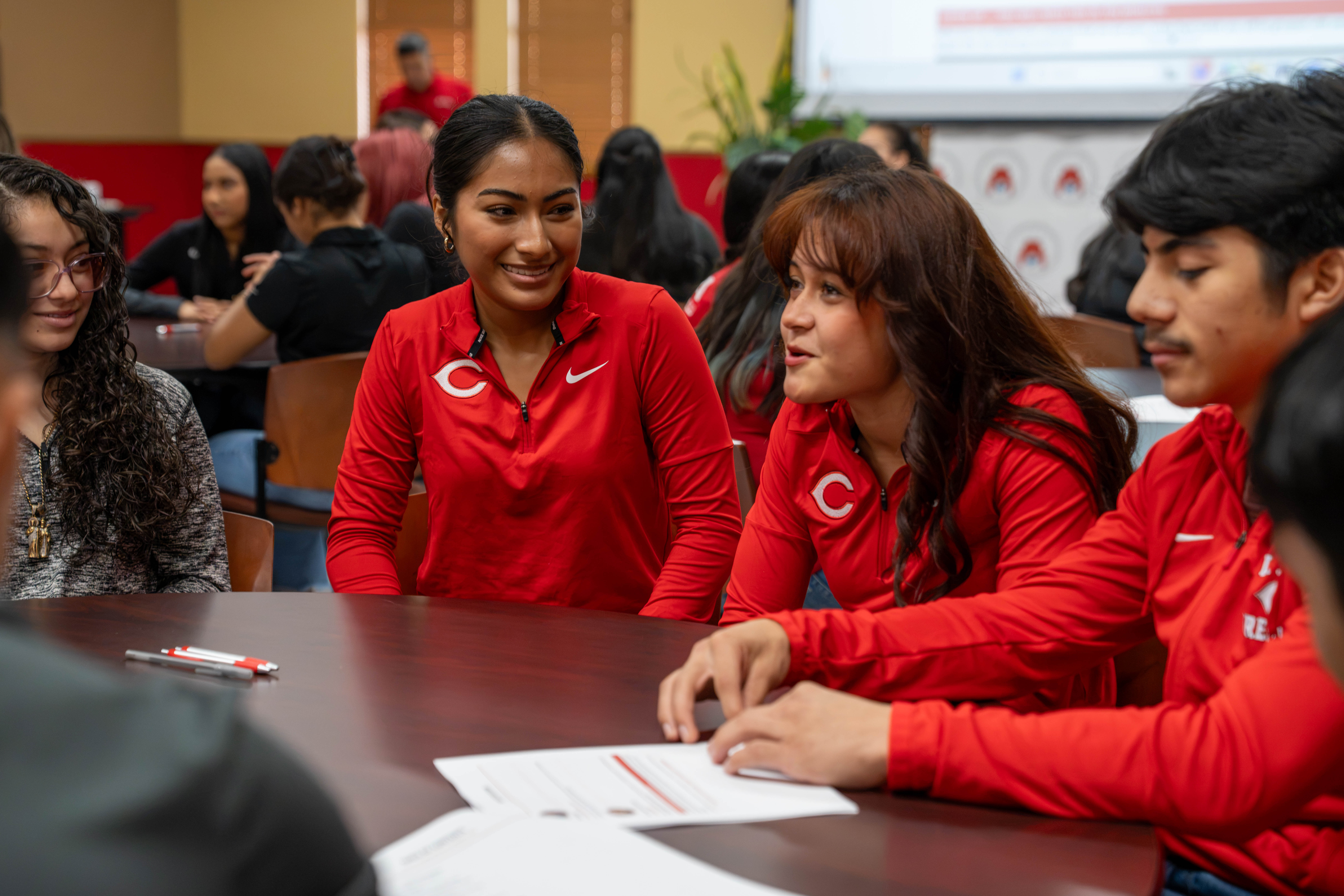 A group of students in matching red jackets sit around a dark wooden table, engaged in conversation during an event.