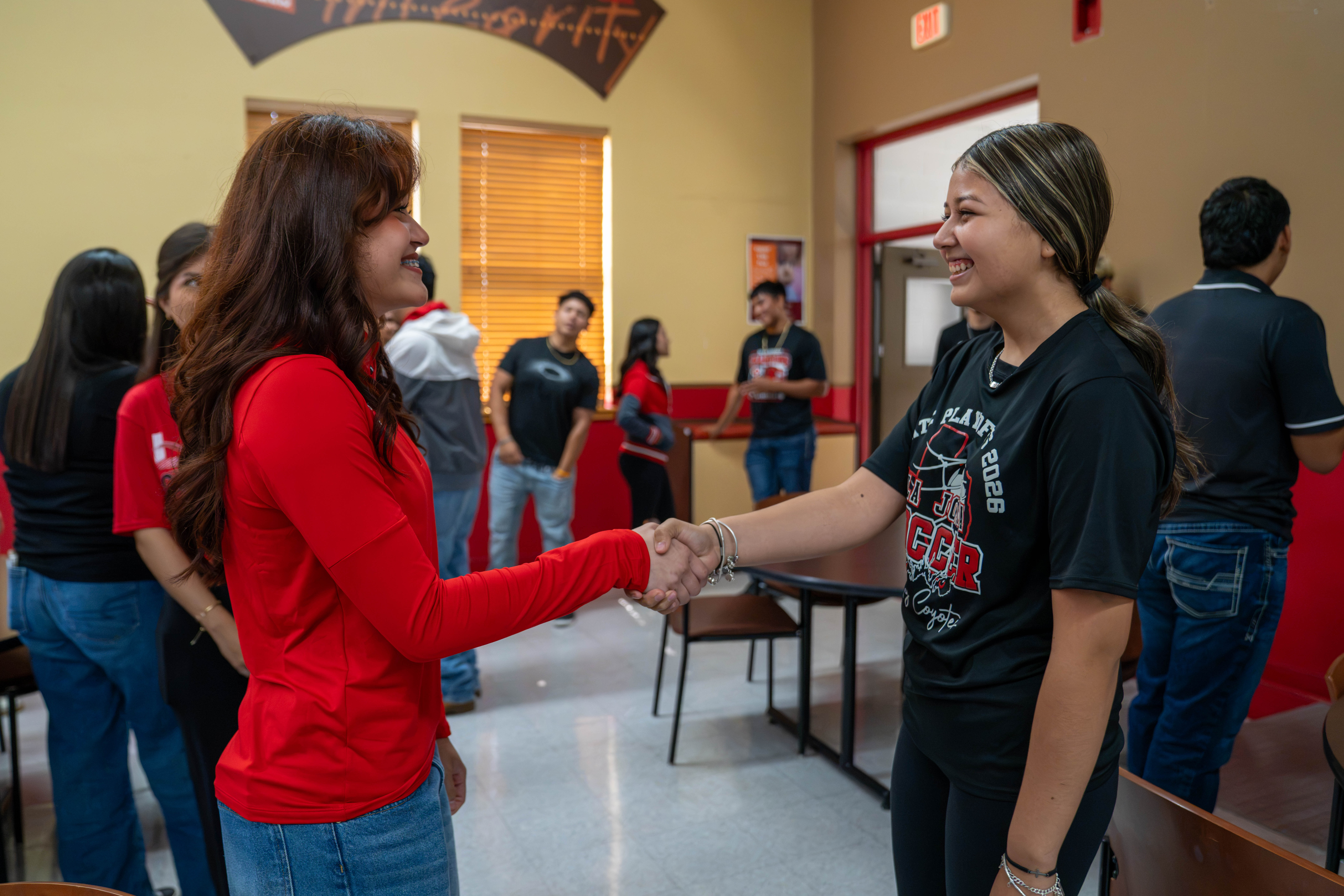 Two smiling young women shake hands indoors, surrounded by peers in a brightly lit room.