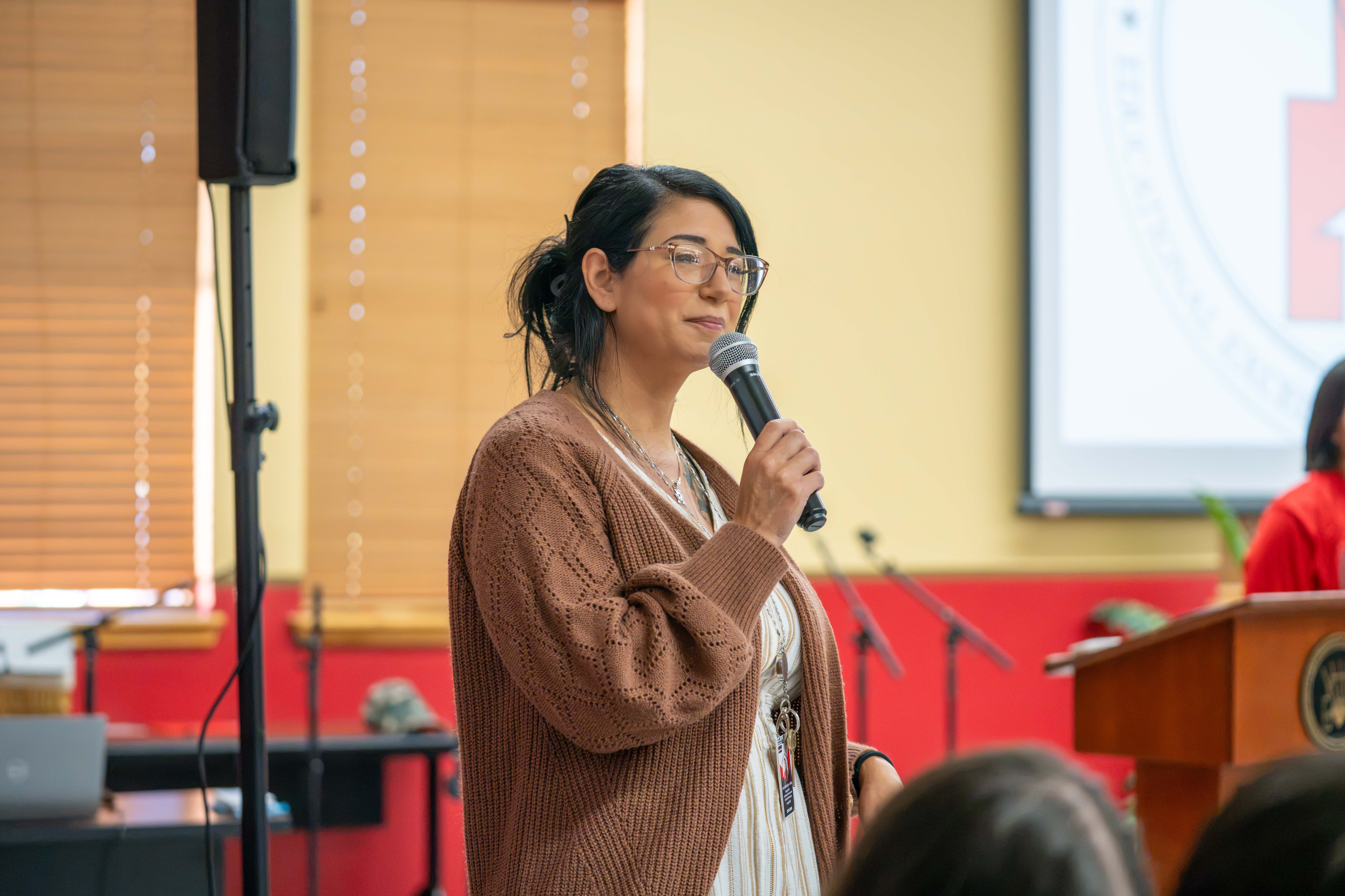 A woman with glasses and dark hair speaks into a microphone while wearing a brown cardigan.