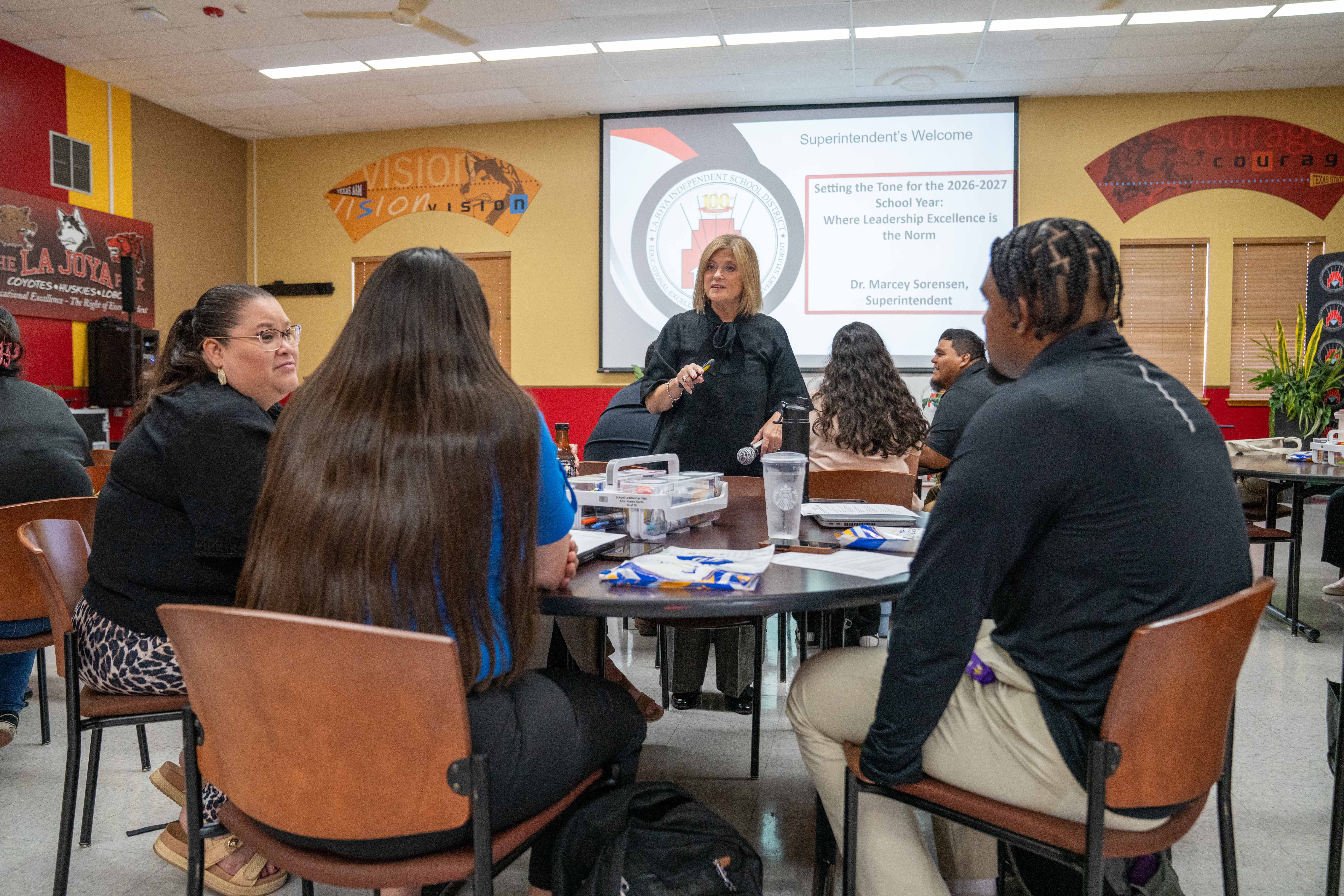 A superintendent presents to a group of people seated around a table in a classroom.