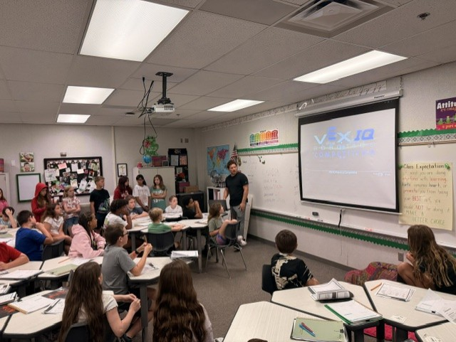 A teacher presents to a classroom of students gathered around desks, with a VEX IQ Competition logo projected on a screen.