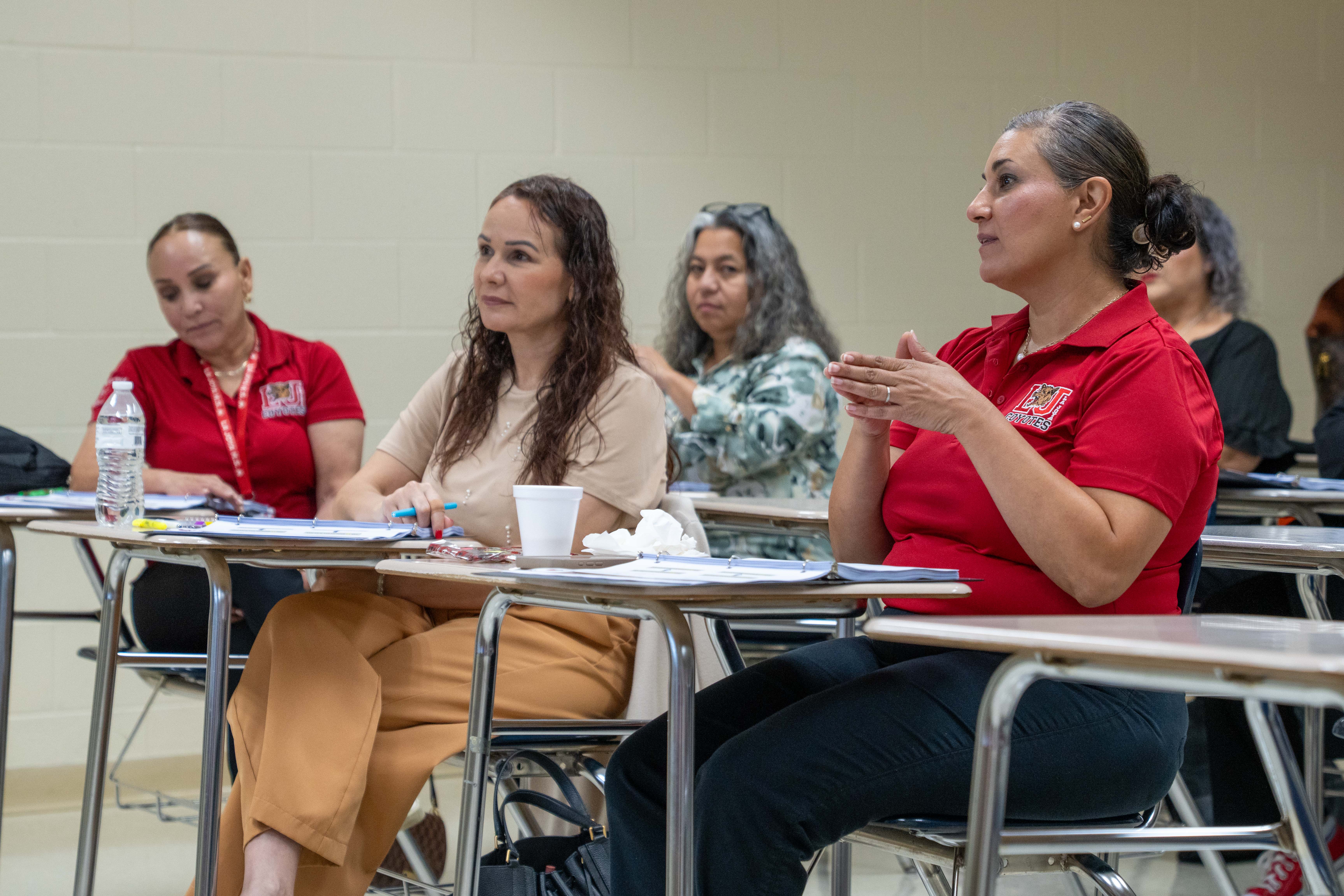 A group of adults attend a class, seated at desks with papers and water bottles.