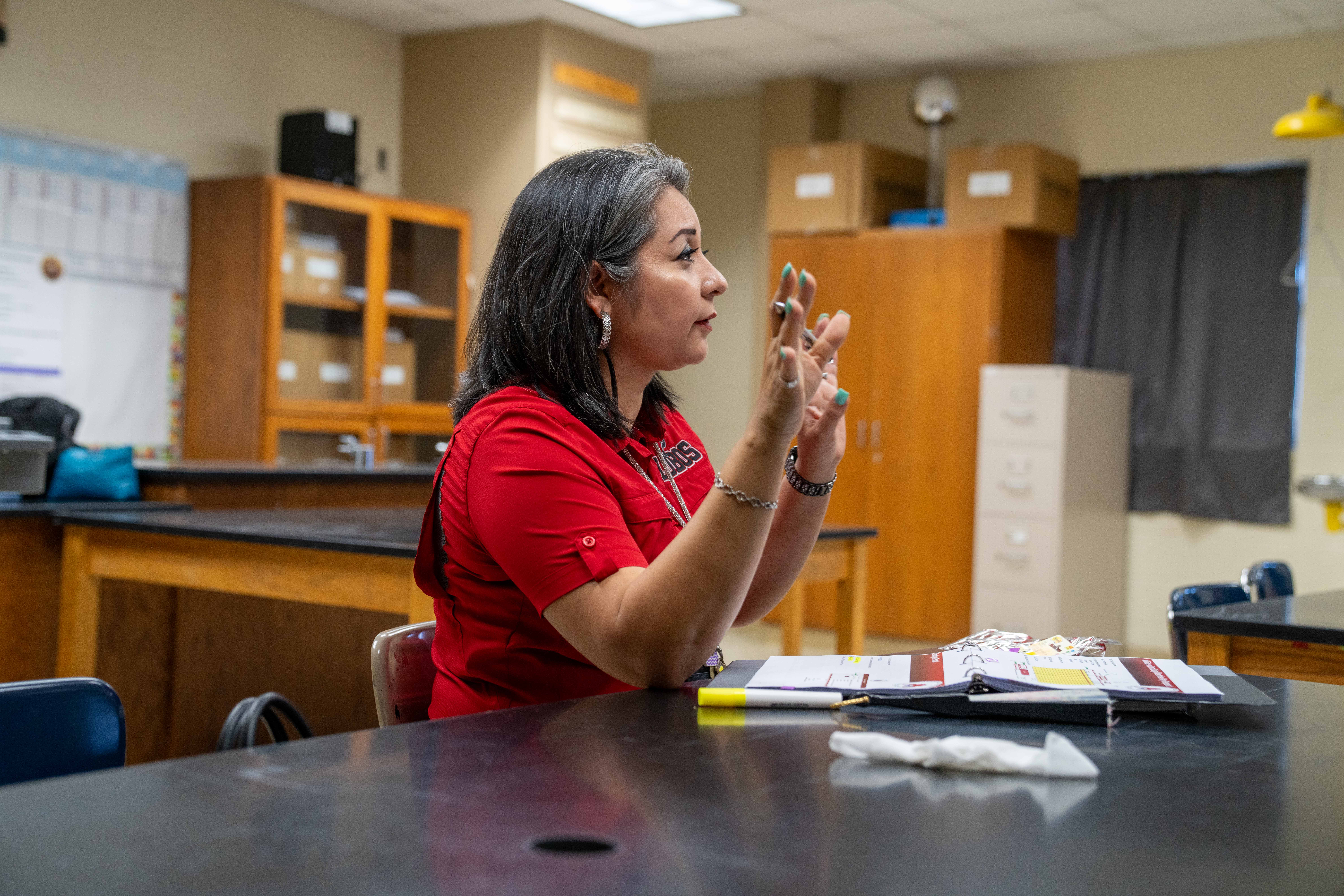 A woman in a red shirt gestures with her hands while sitting at a table in a classroom.